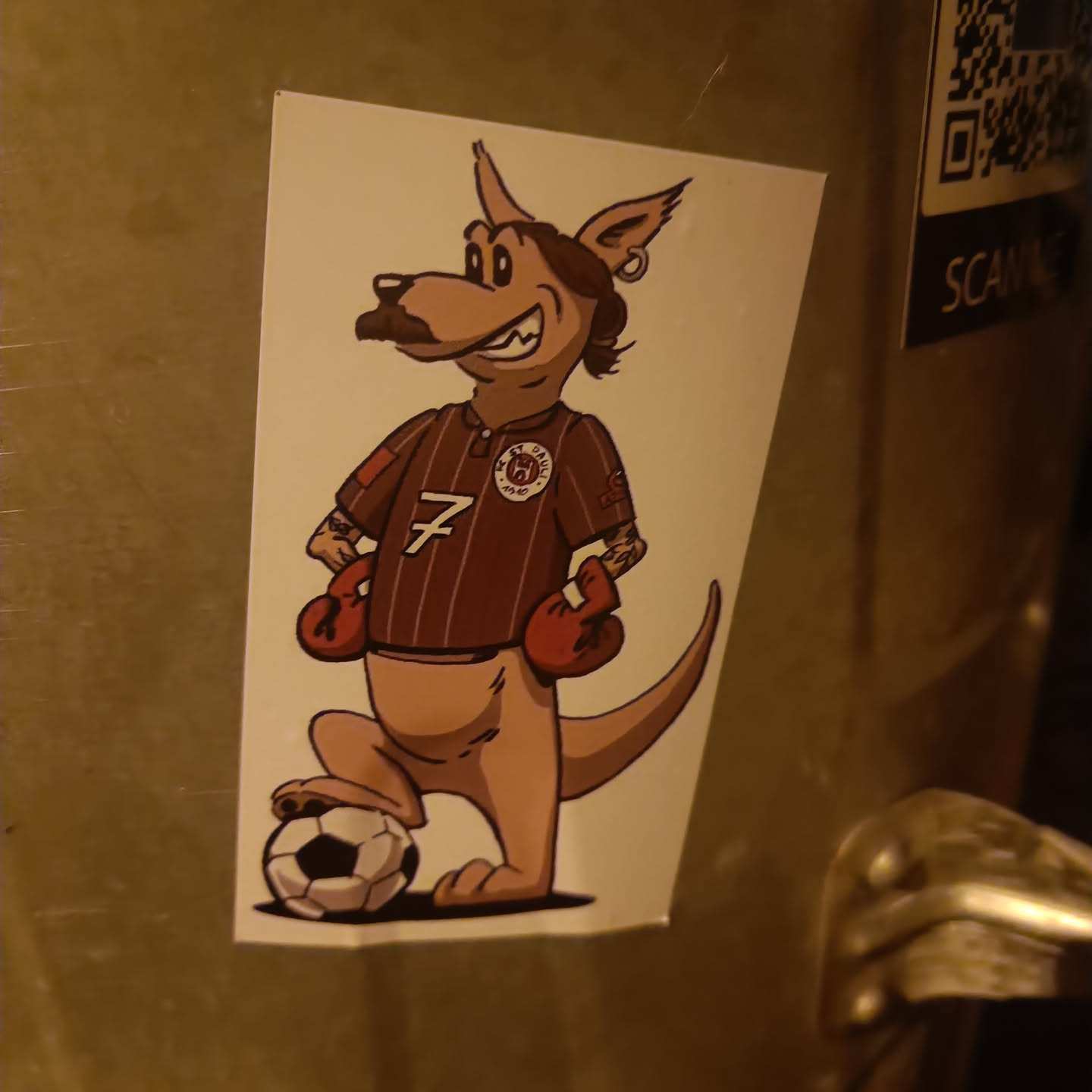 This sticker features a cartoon illustration of a kangaroo wearing a maroon soccer jersey with the number 7 and boxing gloves. The kangaroo is standing with one foot on a soccer ball.  The style is slightly vintage and has a slightly rough texture, reminiscent of a screen print. 