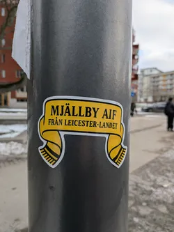The sticker is shaped like a stylized football scarf, predominantly yellow with a thin white border. The top section of the scarf, which forms a banner, features the text 'MJÄLLBY AIF' in bold black capital letters. Below this, on a slightly smaller line, is 'FRÅN LEICESTER-LANDET' also in black capital letters. The two ends of the scarf hang down from the main banner, each featuring black and yellow stripes, mimicking the fringed ends of a traditional football scarf.