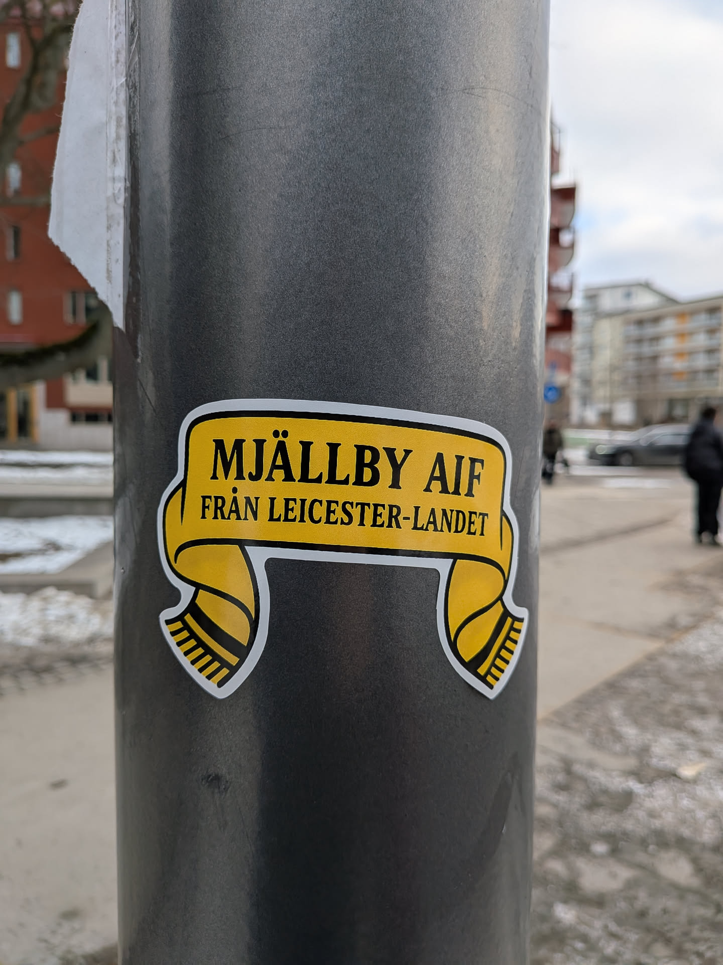 The sticker is shaped like a stylized football scarf, predominantly yellow with a thin white border. The top section of the scarf, which forms a banner, features the text 'MJÄLLBY AIF' in bold black capital letters. Below this, on a slightly smaller line, is 'FRÅN LEICESTER-LANDET' also in black capital letters. The two ends of the scarf hang down from the main banner, each featuring black and yellow stripes, mimicking the fringed ends of a traditional football scarf.