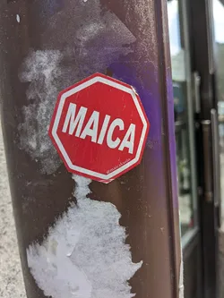 Red octagonal sticker with white text that says MAICA. It has a social media handle on the bottom.  The sticker is affixed to a brown pole.