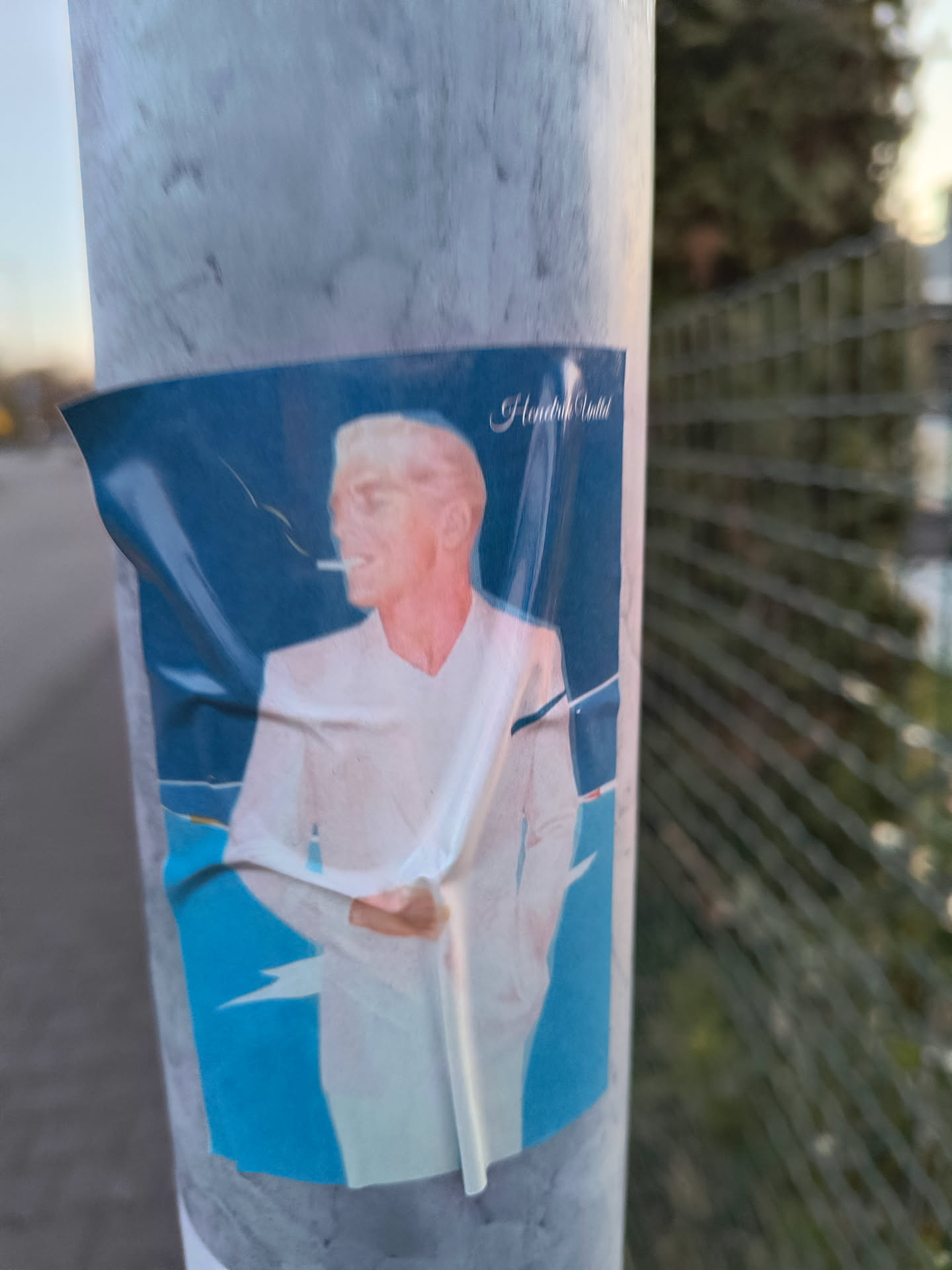 The sticker features a man with white hair and skin, smoking a cigarette. He is wearing a white suit against a blue background. The sticker is partially wrinkled and affixed to a metal pole.