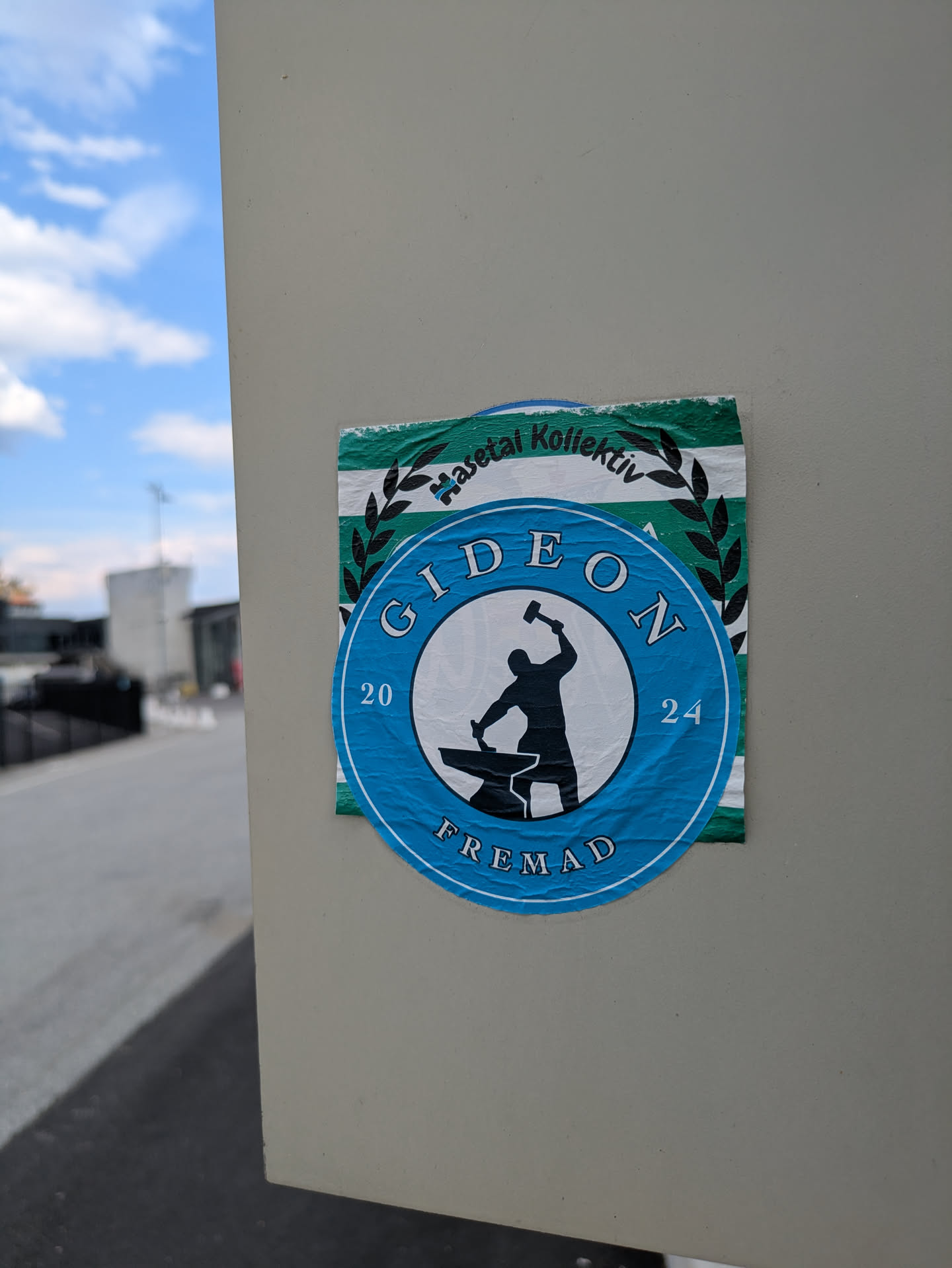 The central sticker is round and predominantly blue with a white inner circle. Inside this circle is a black silhouette of a person, likely a blacksmith, striking an anvil with a hammer. Around the outer blue ring, the word "GIDEON" is written at the top, "FREMAD" at the bottom, and the numbers "20" and "24" are placed on the left and right sides respectively, suggesting a year. Partially visible behind this blue sticker is a rectangular sticker with horizontal green and white stripes and a black laurel wreath design, with text "Haseta Kollektiv" visible.