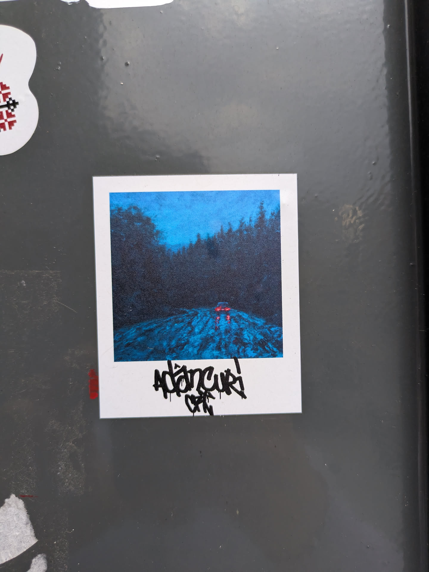 The sticker features a polaroid-style photo of a forest. It appears to be nighttime with a road and trees and two red lights on the road. Below the photo, there is some handwritten text.