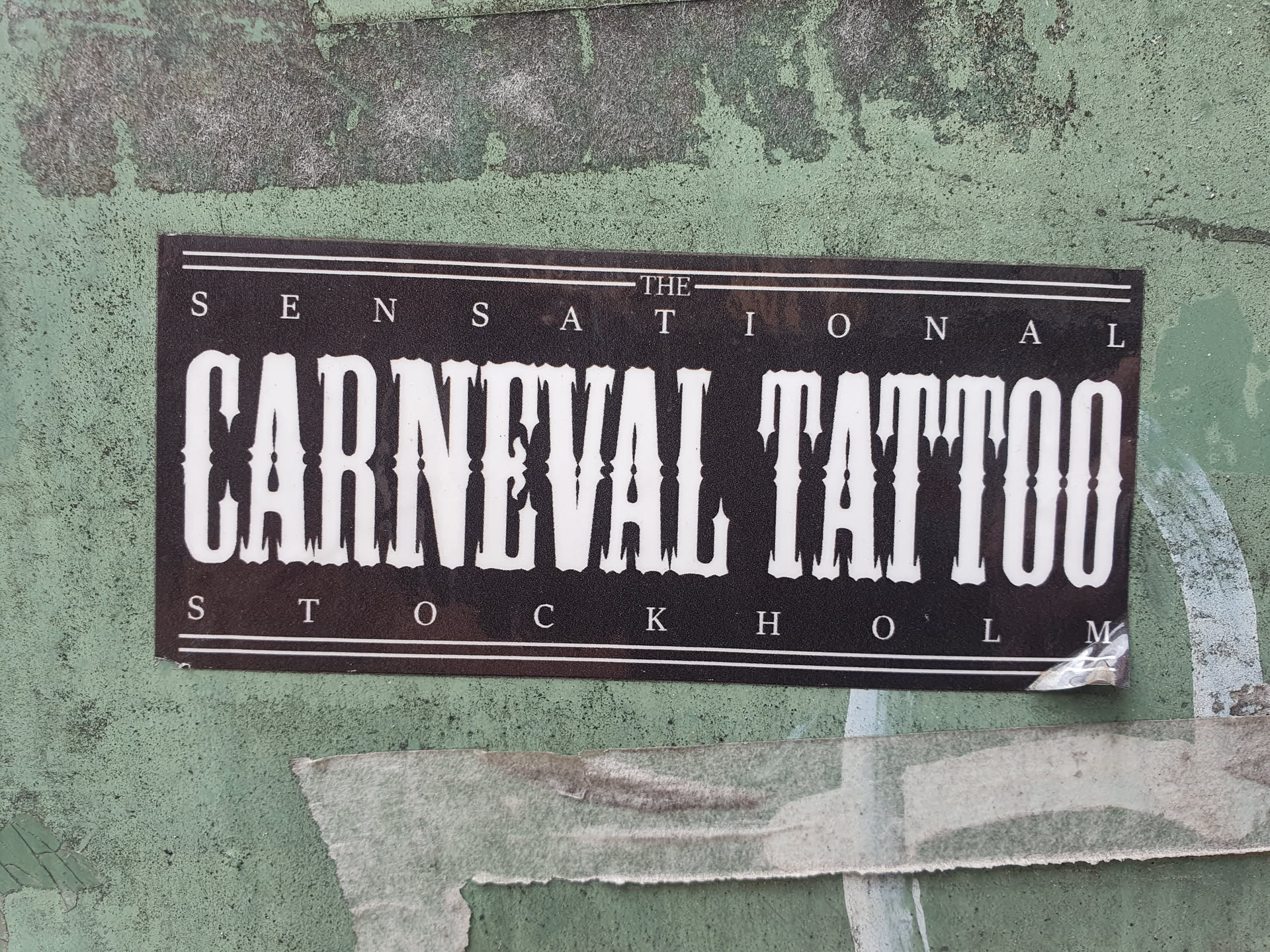 A black and white sticker with the text "The Sensational Carnival Tattoo Stockholm" in a bold, western-style font. The sticker is affixed to a weathered green wall.