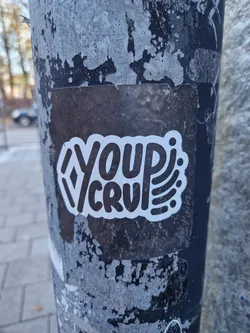 A black and white sticker with the text "Your Cru" in a stylized font. The sticker is on a weathered pole.