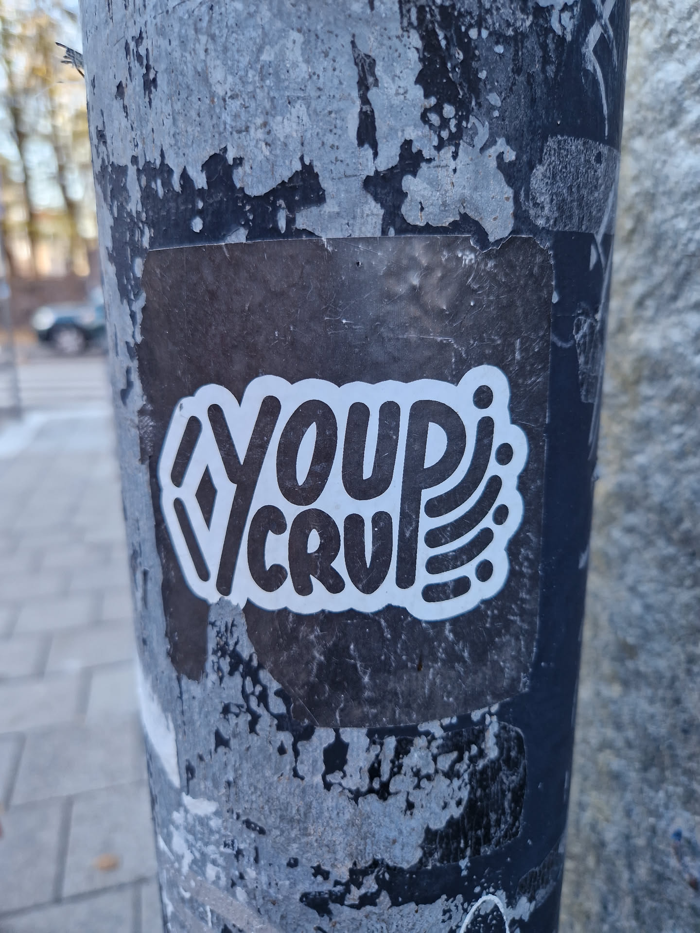 A black and white sticker with the text "Your Cru" in a stylized font. The sticker is on a weathered pole.
