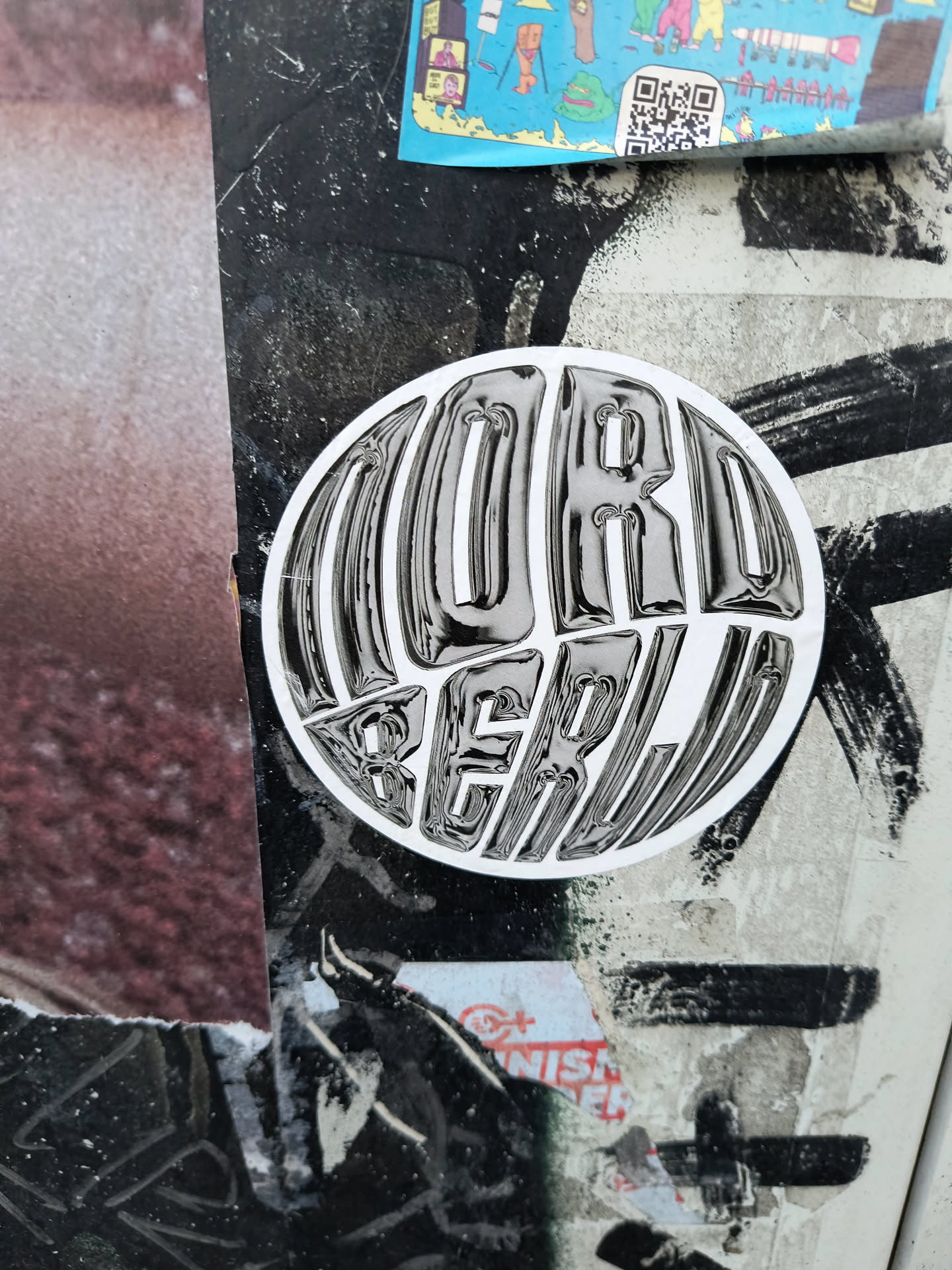 The sticker is circular and features text with a glossy, metallic sheen. The text is rendered in a bold, stylized font, with each letter appearing to have depth and a reflective surface. The colors are monochromatic, primarily black and white, with highlights and shadows that emphasize the three-dimensional effect of the lettering. The sticker is placed on a surface with other layered posters and graffiti.