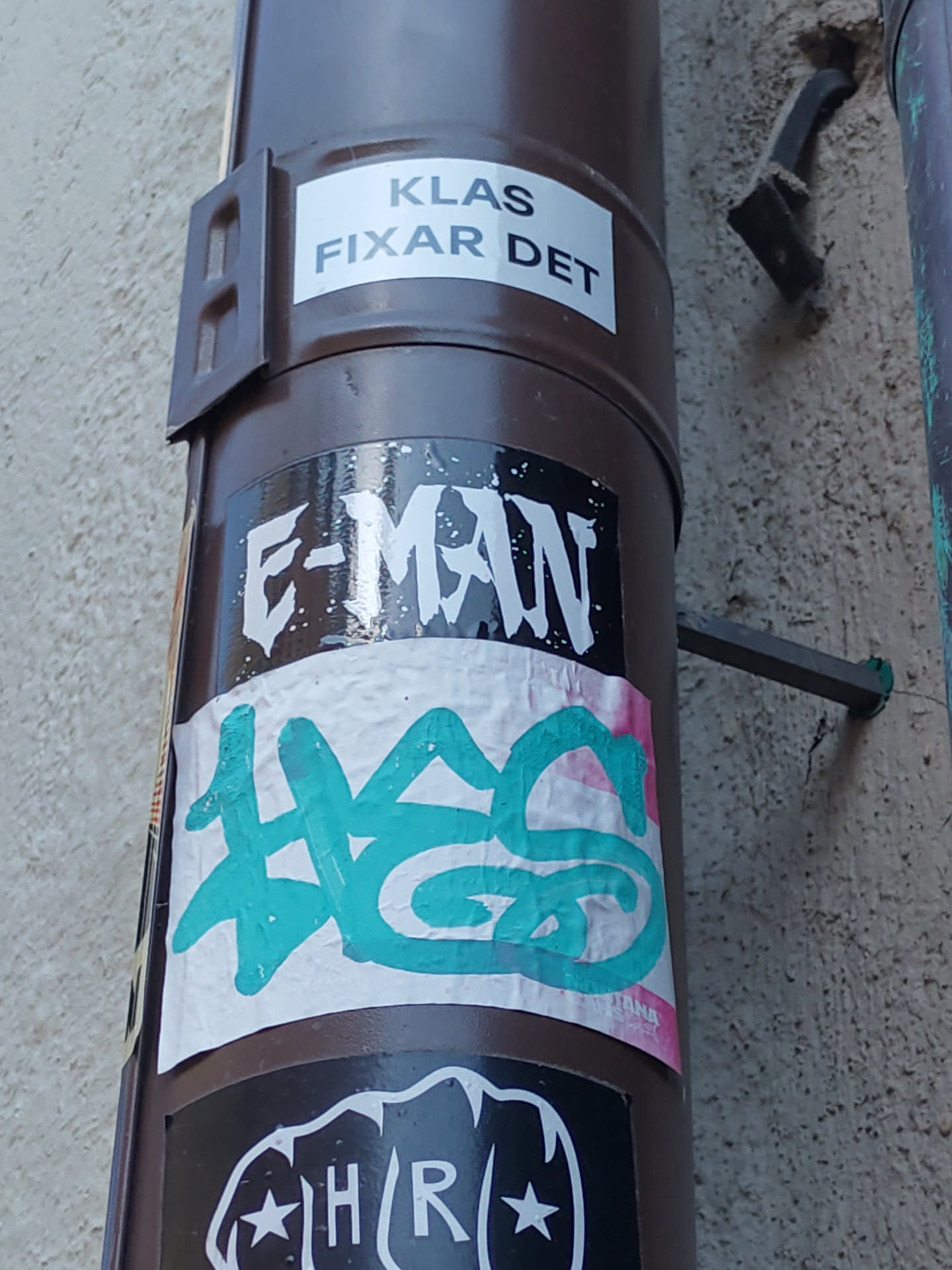 A teal and white graffiti sticker with illegible text is placed on a brown pipe.  Above it is a black sticker with the words "E-MAN" in white. There is also a white sticker above that that says "KLAS FIXAR DET". Below it is a black sticker with a fist and the text "HR" .