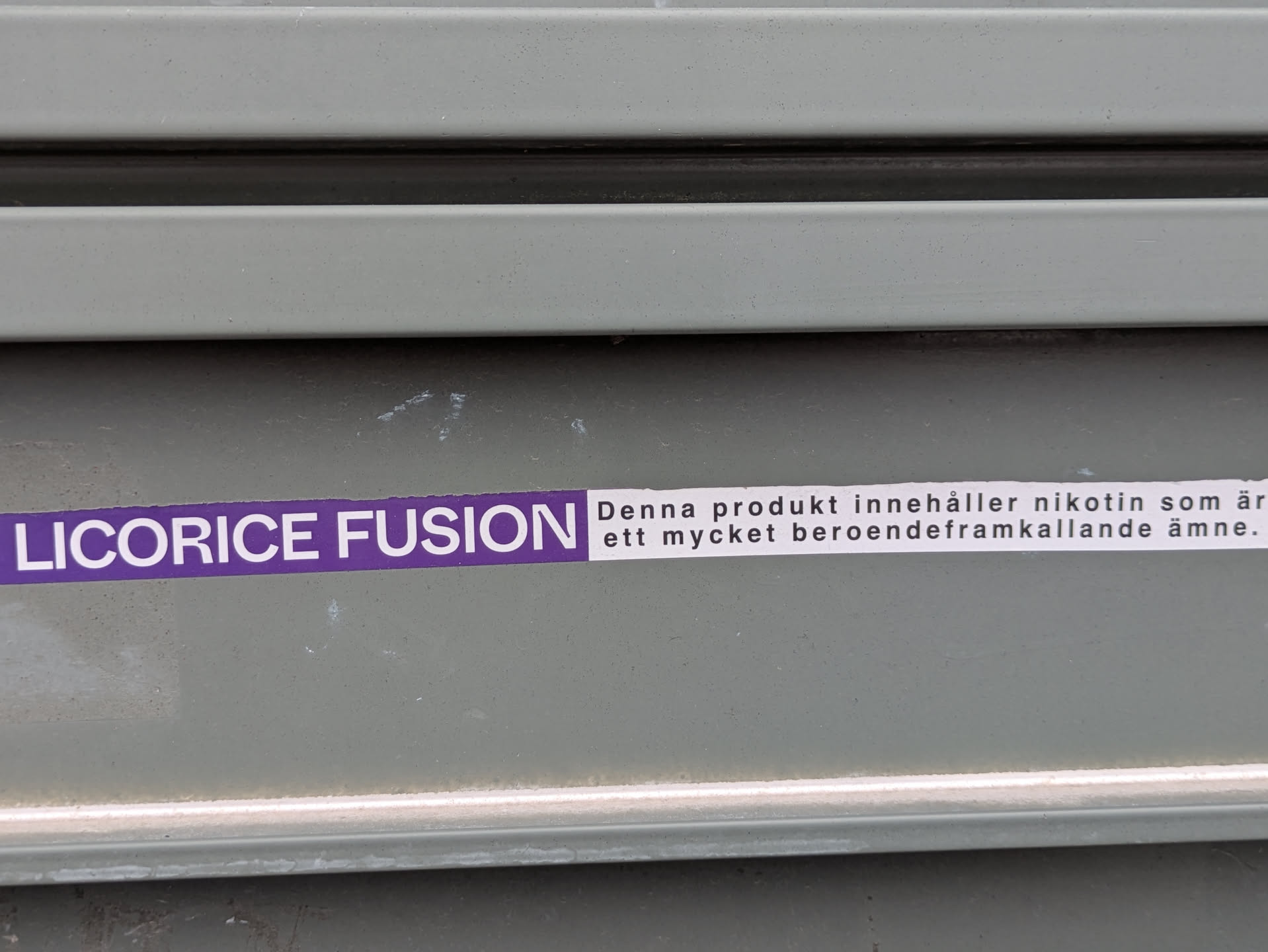 The image shows a sticker on a grey corrugated surface. The sticker is horizontally oriented, with the text "LICORICE FUSION" in purple on the left side, followed by a white label with the text "Denna produkt innehåller nikotin som är ett mycket beroendeframkallande ämne."