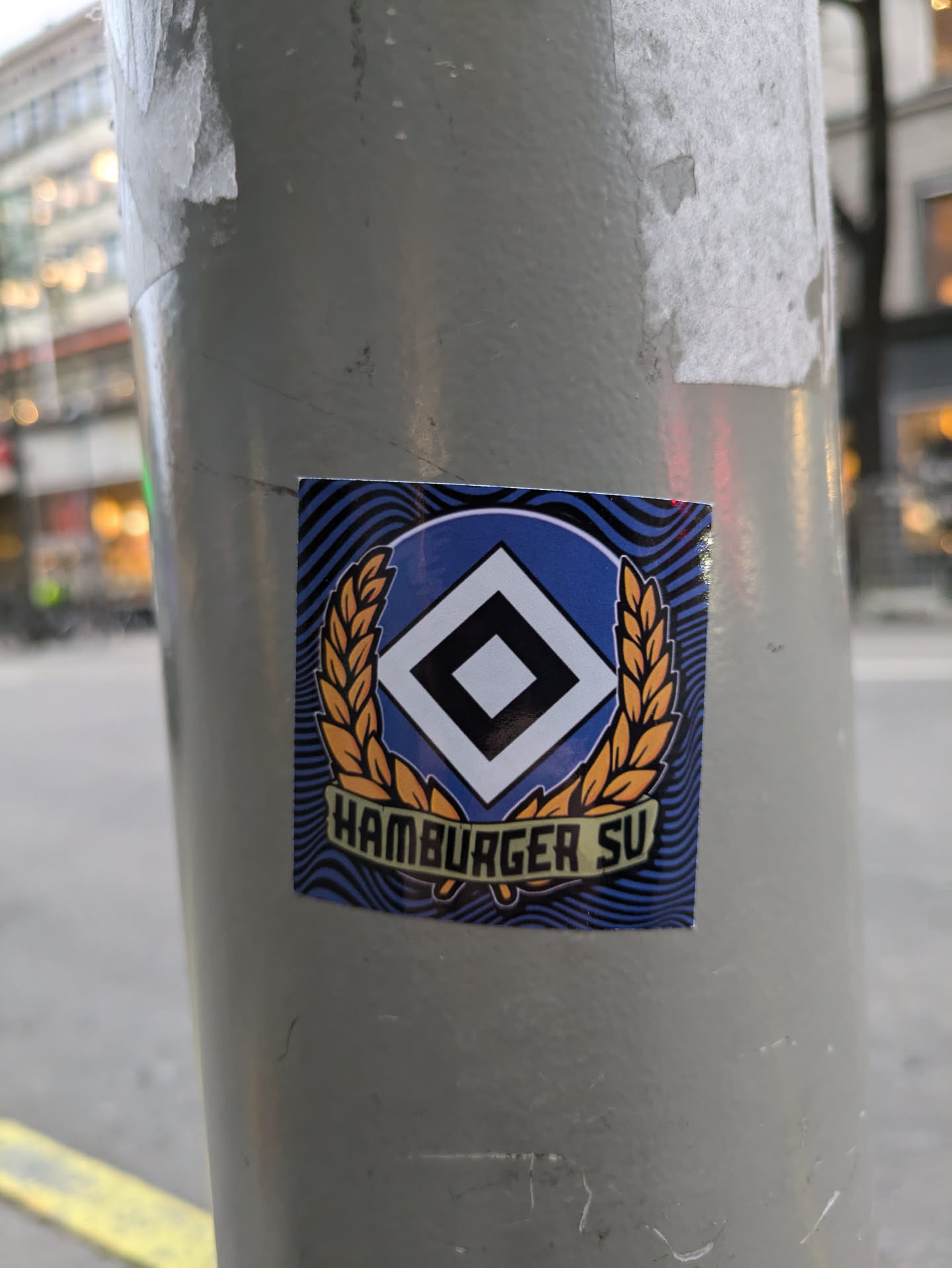 A sticker featuring the logo of Hamburger SV, a German football club. The logo is a stylized diamond shape with a wreath. The text "Hamburger SV" is visible at the bottom of the sticker. The sticker has a wavy blue background.