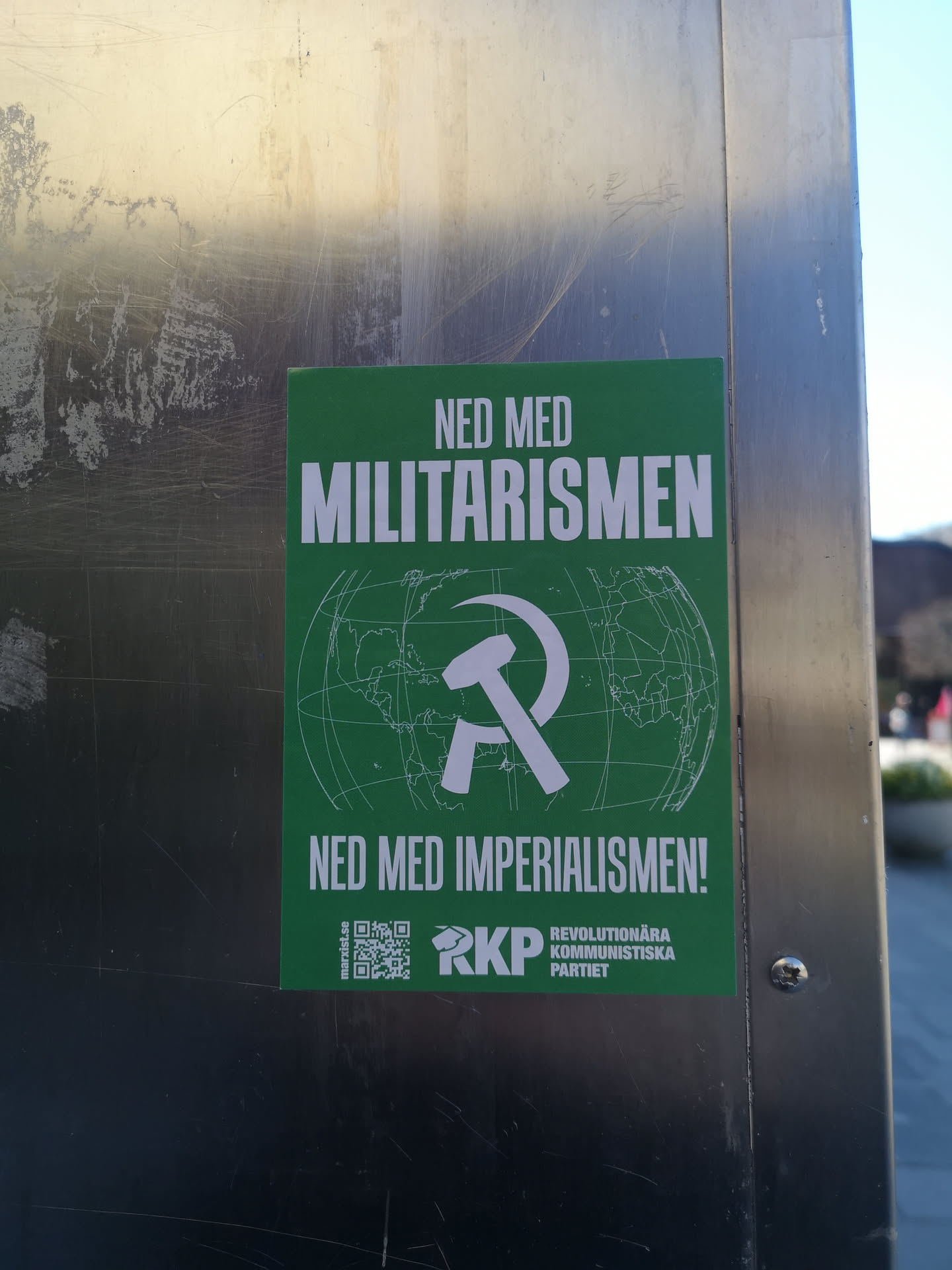 A rectangular green sticker featuring bold white text at the top and bottom. The top text reads 'NED MED MILITARISMEN' (Down with Militarism) and the bottom text reads 'NED MED IMPERIALISMEN!' (Down with Imperialism!). In the center, a prominent white hammer and sickle symbol is overlaid on a faint white outline of a globe, depicting continents and grid lines. At the bottom left, a small QR code is present, next to the text 'marxist.se'. To the right of the QR code, the white acronym 'RKP' is displayed, followed by the full name 'REVOLUTIONÄRA KOMMUNISTISKA PARTIET' (Revolutionary Communist Party) in smaller white text.