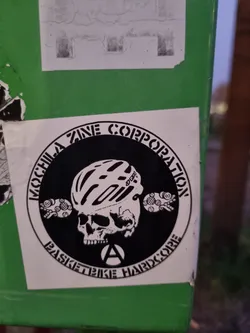 Black and white sticker with a skull wearing a bicycle helmet. The text around the circle says "Mochila Zine Corporation Basketbike Hardcore".