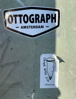 A sticker with a drawing of a test tube and the text "Test your drugs". The sticker is white with black ink.  It's affixed to a grey surface.  There is a smaller sticker above it that reads "OTTOGRAPH - AMSTERDAM -"