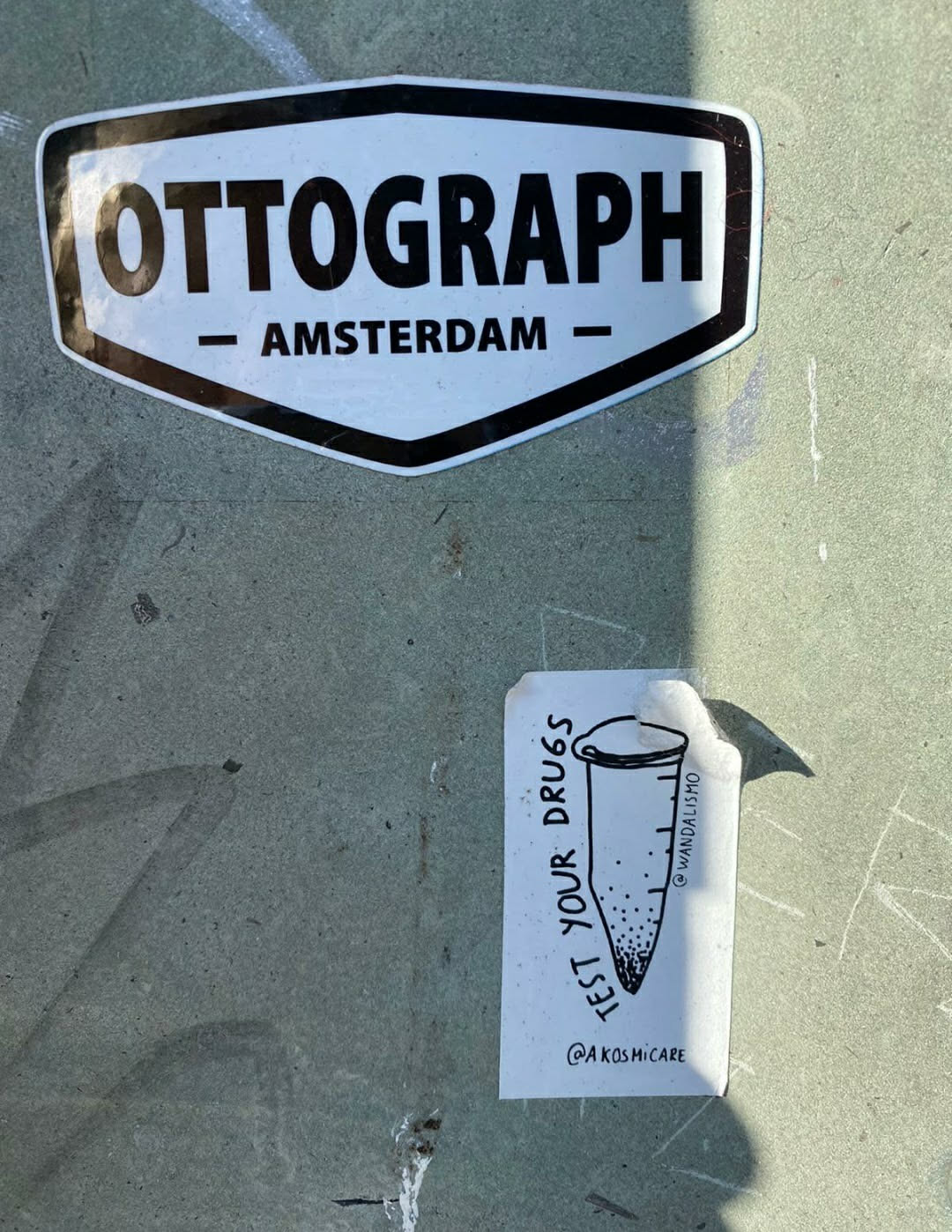A sticker with a drawing of a test tube and the text "Test your drugs". The sticker is white with black ink.  It's affixed to a grey surface.  There is a smaller sticker above it that reads "OTTOGRAPH - AMSTERDAM -"