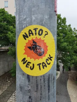A yellow round sticker with a black and orange illustration of a bomb with wings. The text NATO? Nej tack is visible on the sticker. Nej tack is Swedish for No thanks. 