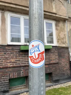 A circular sticker featuring the F.C. Hansa Rostock club crest. It has a white background with a blue outer border. The upper half displays a stylized blue ship with a white sail, on which a white lion rampant is depicted. A small red flag or pennant flies from the ship's mast. The lower half features a curved red banner-like element with the white text 'F.C. HANSA' written along its curve. A red anchor-like symbol or stylized cross is positioned between the ship and the text. The primary colors are white, blue, and red.