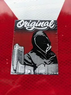 A sticker featuring a hooded figure in a grayscale style, set against a red background.  The figure is positioned in front of a cityscape. The word "Original" is written in a stylized script font at the top of the sticker.