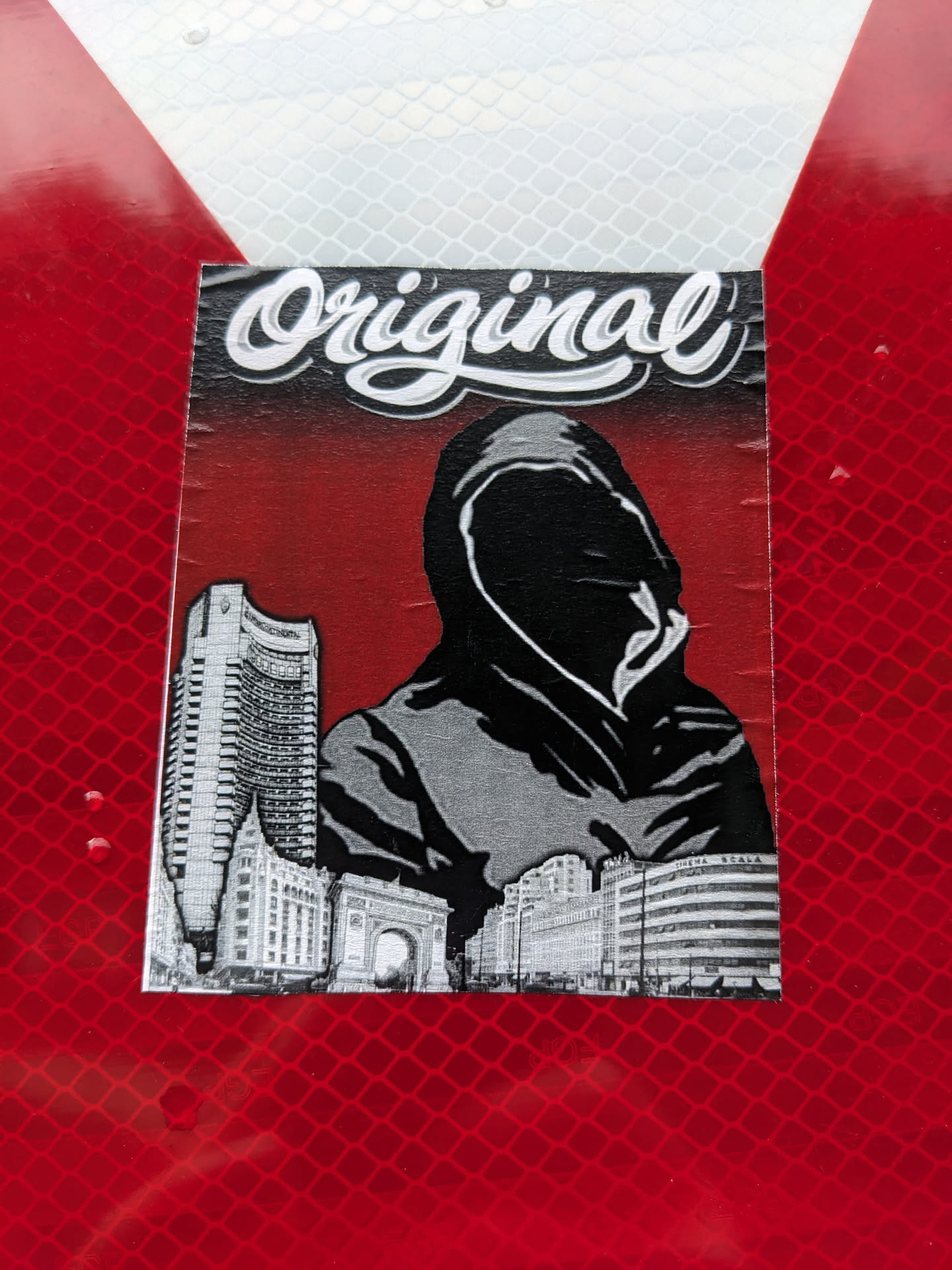 A sticker featuring a hooded figure in a grayscale style, set against a red background.  The figure is positioned in front of a cityscape. The word "Original" is written in a stylized script font at the top of the sticker.