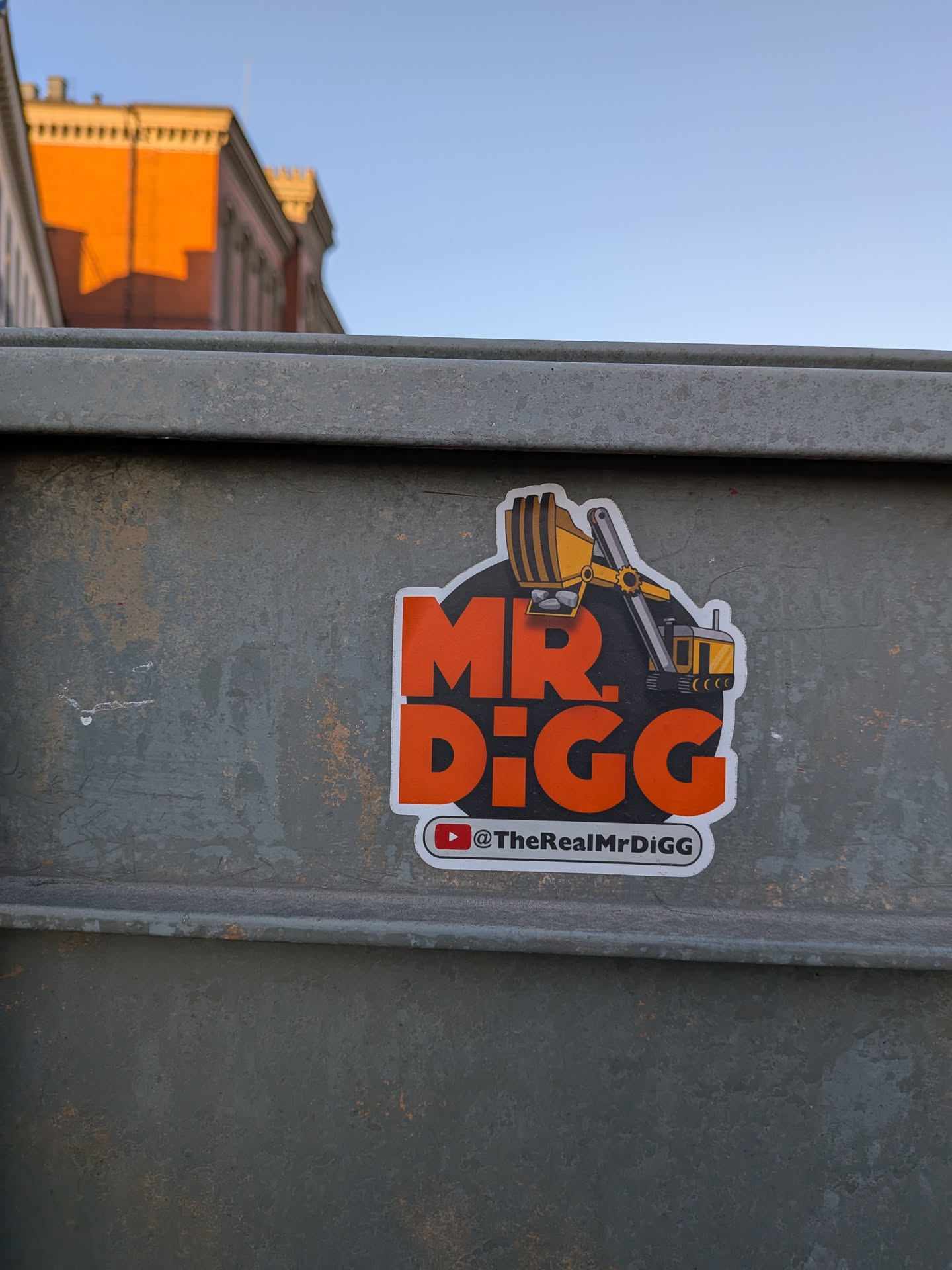 The sticker features a bold, orange 'MR. DIGG' text against a dark grey, almost black, irregular circular background with a white outline. An illustration of a yellow and black excavator is positioned above and to the right of the text, with its bucket extended over the 'MR.' part of the text, appearing to be 'digging' into it. Below the main text, there's a smaller white rectangular strip with a red YouTube play button logo and the handle '@TheRealMrDiGG' in black text.