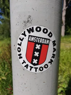 Round sticker featuring the text 'Hollywood Tattoo' arranged circularly around a central emblem. The emblem consists of a red and black shield with three white crosses, with the word 'Amsterdam' written above it. The sticker is affixed to a grey metal pole.