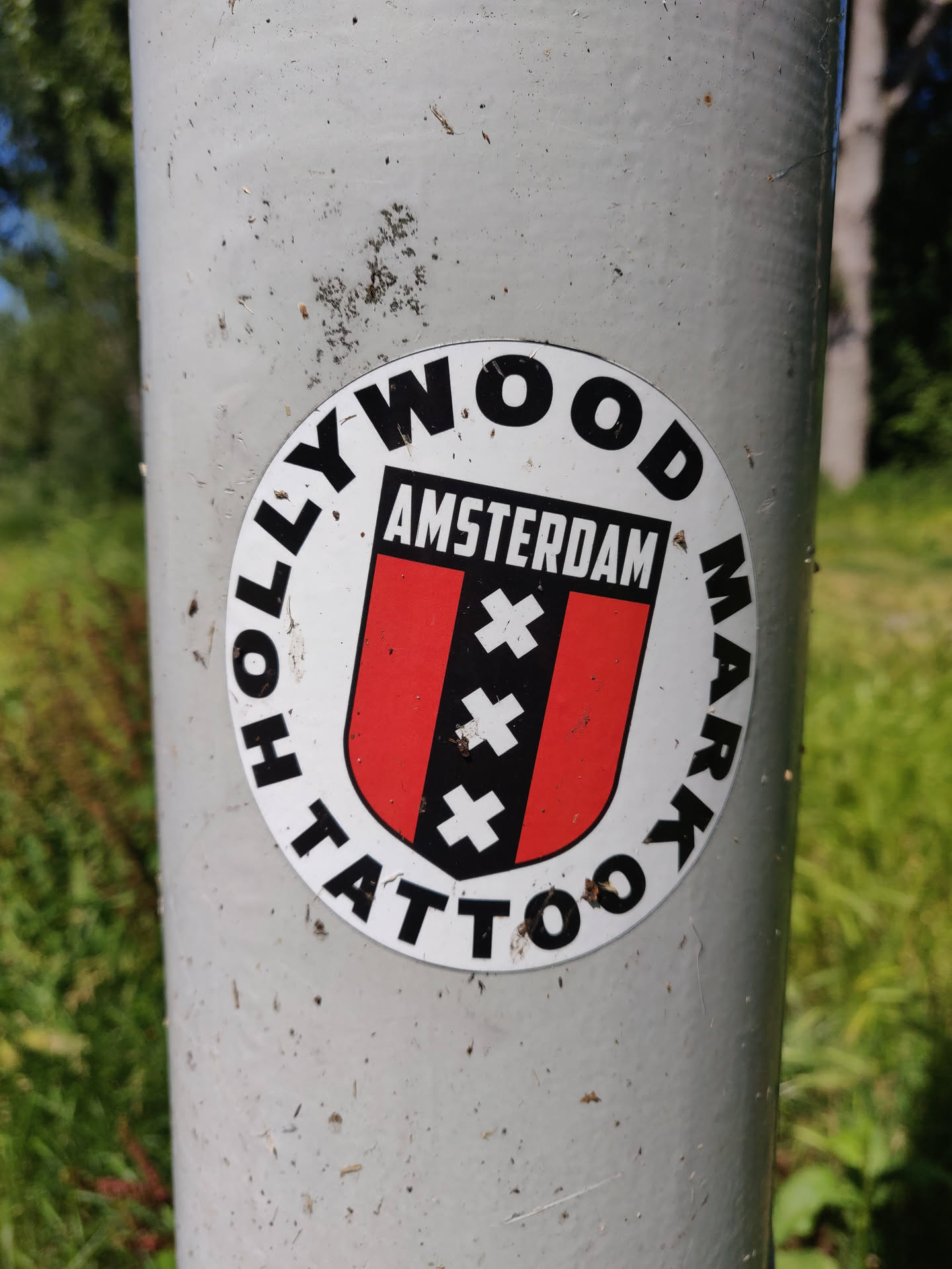Round sticker featuring the text 'Hollywood Tattoo' arranged circularly around a central emblem. The emblem consists of a red and black shield with three white crosses, with the word 'Amsterdam' written above it. The sticker is affixed to a grey metal pole.
