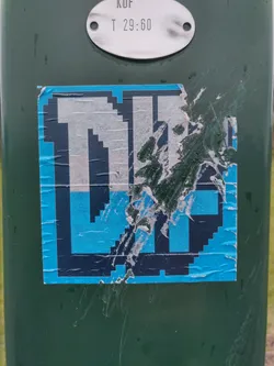 A partially damaged sticker featuring pixel art. The design is primarily light blue and black, forming a blocky letter or symbol. The sticker is affixed to a dark green pole. 