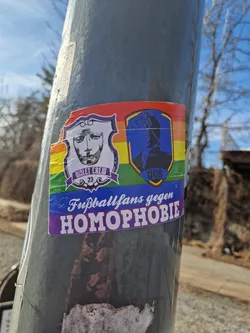 Sticker expressing the message of football fans against homophobia. It features two logos, one with a face and the text "Violet Crew 23", and another with a figure and the letters "UCS".  The sticker uses rainbow colors as a background.