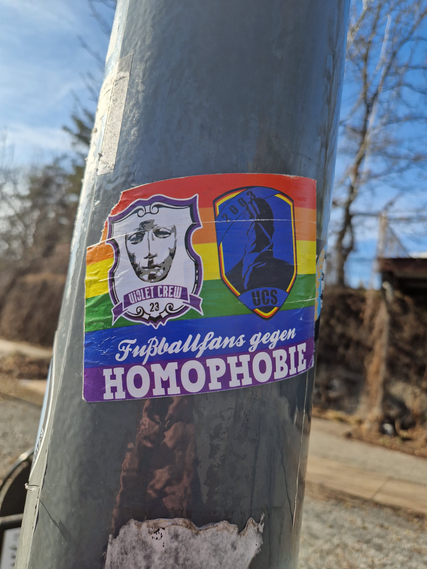 Sticker expressing the message of football fans against homophobia. It features two logos, one with a face and the text "Violet Crew 23", and another with a figure and the letters "UCS".  The sticker uses rainbow colors as a background.
