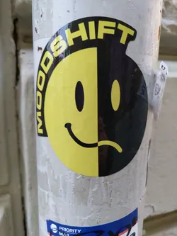 A sticker featuring a split smiley face, half happy and half sad, with the word "Moodshift" circling it. The sticker is placed on a white pole.
