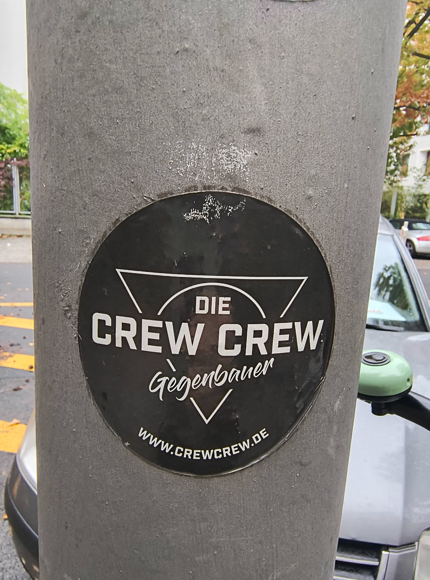 The sticker is circular and predominantly black with white text and graphics. At the top, a white triangle points downwards. Below this, the words 'DIE CREW CREW' are prominently displayed in bold, sans-serif white capital letters. Beneath 'CREW CREW', the name 'Gegenbauer' is written in a flowing, cursive white font. At the bottom of the circle, the website 'WWW.CREWCREW.DE' is written in white, sans-serif capital letters. The sticker appears to be slightly worn or aged, with some minor scuffs and imperfections on its surface.
