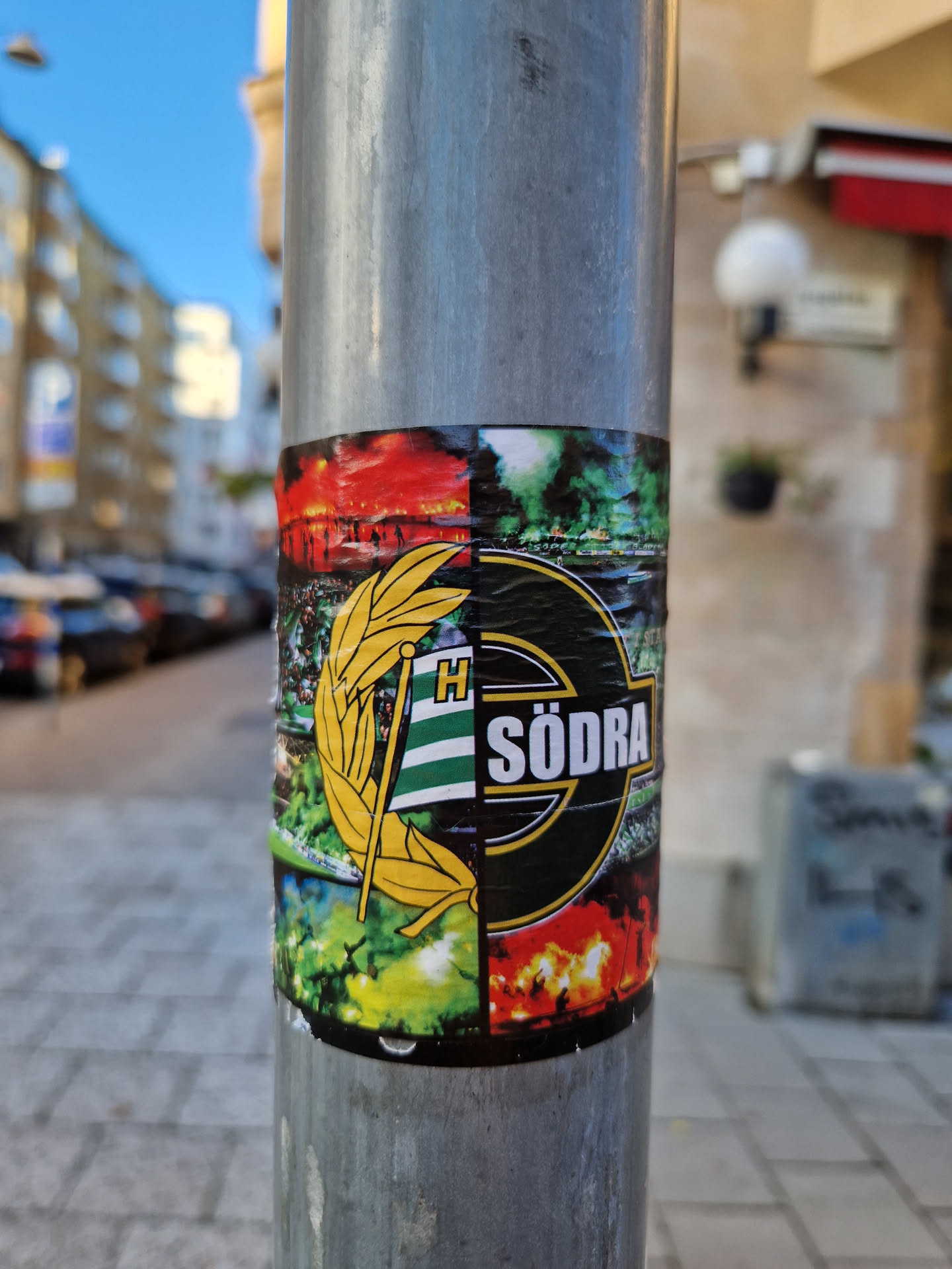 A colorful sticker featuring the logo of what appears to be a Swedish football club, Hödra Södra. The sticker is divided into sections, each with a different graphic element.  It's stuck to a metal pole on a city street. 