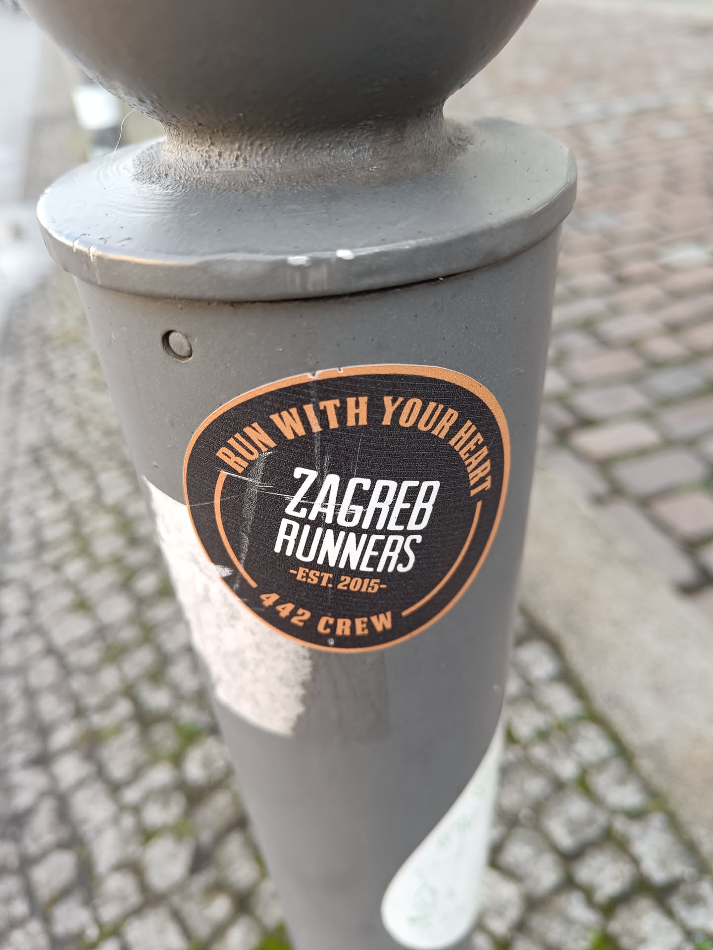 A round black and orange sticker on a grey pole.  The text reads: 'Run with your heart' in a circle around the main text: 'Zagreb Runners -Est. 2015- 442 Crew'.