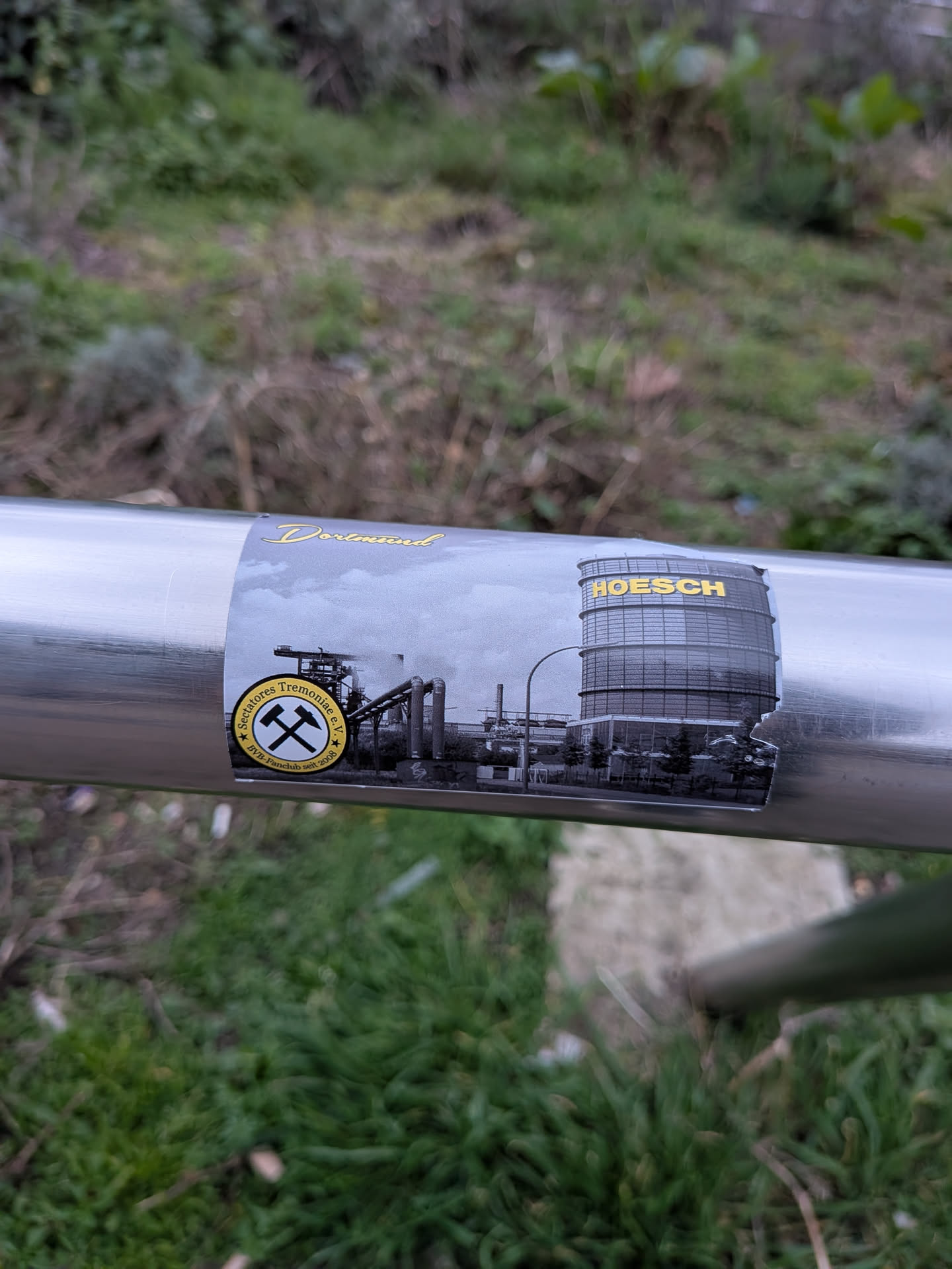 The sticker is a rectangular design applied to a metallic pole, featuring a grayscale depiction of an industrial complex, likely a steel plant, with multiple buildings, smokestacks, and intricate pipework under a cloudy sky. The word "HOESCH" is prominently displayed in gold capital letters on a large cylindrical structure within the industrial scene on the right. Above the main image, the word "Dortmund" is written in an elegant golden script font. In the bottom-left corner, there is a circular logo with a black background, showcasing crossed yellow hammer and pick tools. This logo is encircled by yellow text that reads "Sectatores Tremoniae e.V." at the top and "BVB-Fanclub seit 2008" at the bottom. The sticker shows signs of wear, with a noticeable tear or peeling on its right edge.