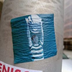 A square sticker featuring a stylized illustration of a girl wearing a hat, rendered in shades of teal and white. The image has a slightly textured or grainy appearance. The sticker is affixed to a gray pole.