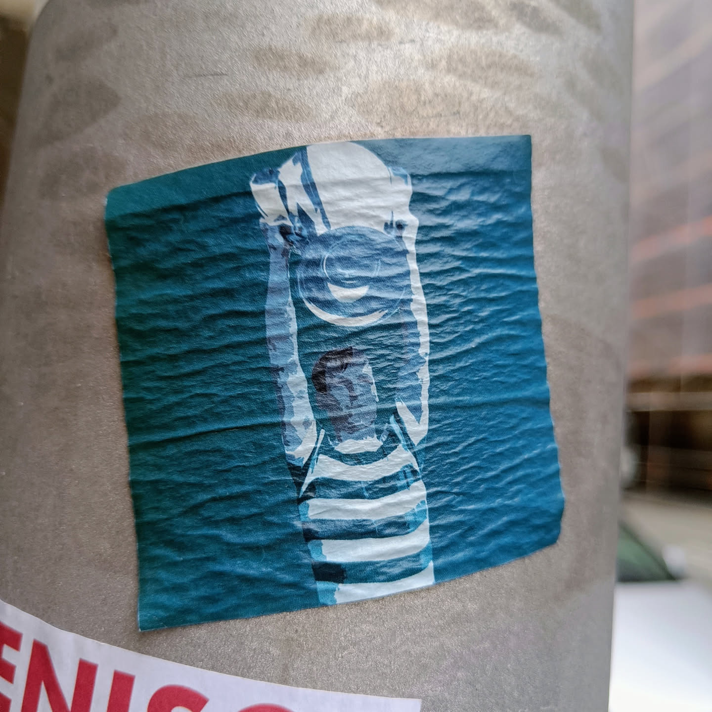A square sticker featuring a stylized illustration of a girl wearing a hat, rendered in shades of teal and white. The image has a slightly textured or grainy appearance. The sticker is affixed to a gray pole.