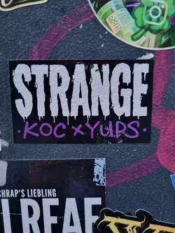 A black sticker with the word "STRANGE" in large, white, stylized letters. Below it, in smaller purple letters, it says ".koc x yups."