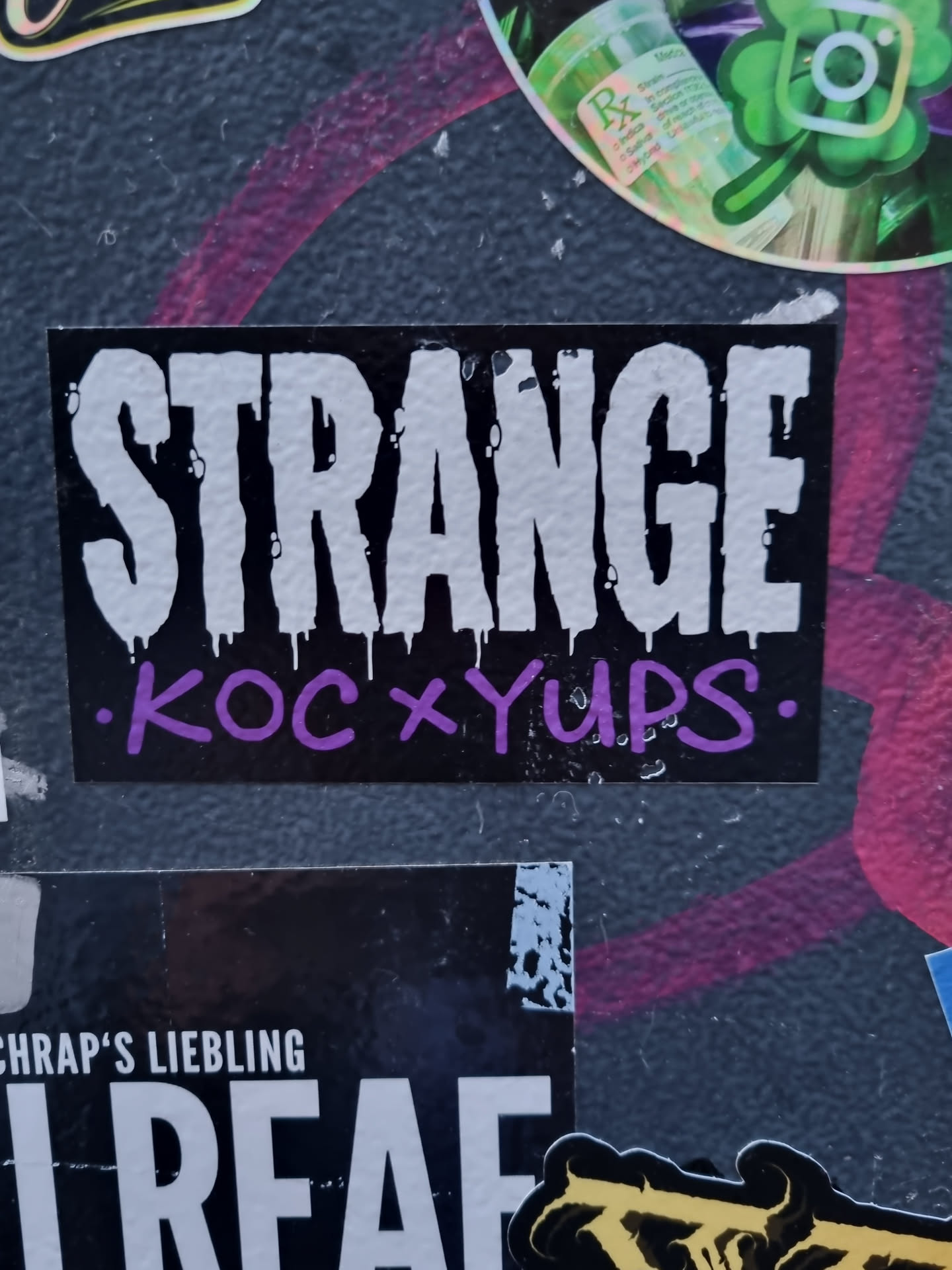 A black sticker with the word "STRANGE" in large, white, stylized letters. Below it, in smaller purple letters, it says ".koc x yups."