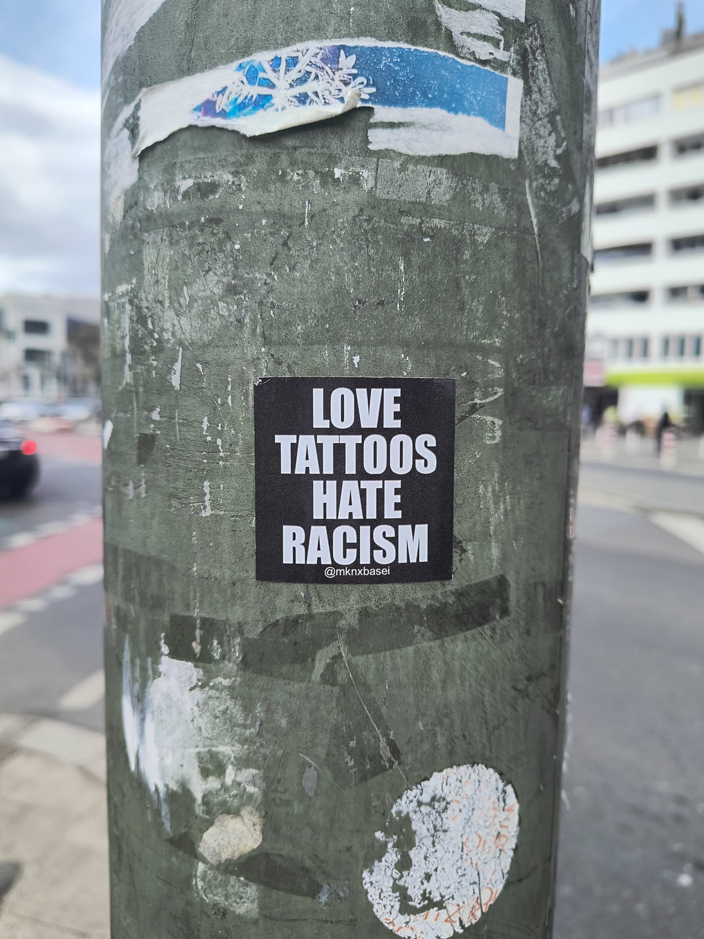 This is a black sticker on a street pole. The sticker says "LOVE TATTOOS HATE RACISM". Below this is the username @mikrobasel.