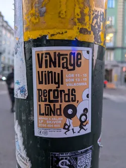 The sticker is predominantly orange with bold black text and stylized illustrations. The top reads 'VINTAGE VINYL' in large, blocky letters. Below this, 'Records, LUND' is written in a similar font. To the right of the text, there are opening hours listed: 'LÖR 11 - 15' and 'SÖN 13 - 15 VÄLKOMNA'. The bottom section of the sticker details the store's offerings: 'KÖPER & SÄLJER LP - SKIVOR' followed by a phone number '0705 404 405'. Further down, the address 'LILLA FISKAREGATAN 4, IN PÅ GÅRDEN' and a phone number '+46 (0) 705 - 404 405' are present. Social media icons for Facebook and Instagram are visible, along with the phone number again. The illustrations depict two cartoonish figures with large eyes, one resembling a vinyl record and the other a small, stick-like character, both in motion. The background of the sticker is a light orange hue.