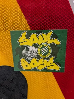 A rectangular sticker with a dark green background, subtly textured with darker splatters or shadows. The main design features the words "SOUL" at the top and "BOYS" at the bottom, rendered in bold, yellow bubble letters with a white outline and a black drop shadow. Centered between the words is a cartoonish, grotesque skull-like creature in white with black outlines and details, characterized by a single large eye, prominent teeth, and a distorted facial structure. To the right of the skull is a circular emblem with a yellow laurel wreath border. Inside the emblem, on a dark green background, is a white flag with horizontal stripes, depicted on a pole. There appears to be small, indistinct text on the flag. The sticker itself shows visible creases and slight wear, indicating it's a physical object applied to a surface.