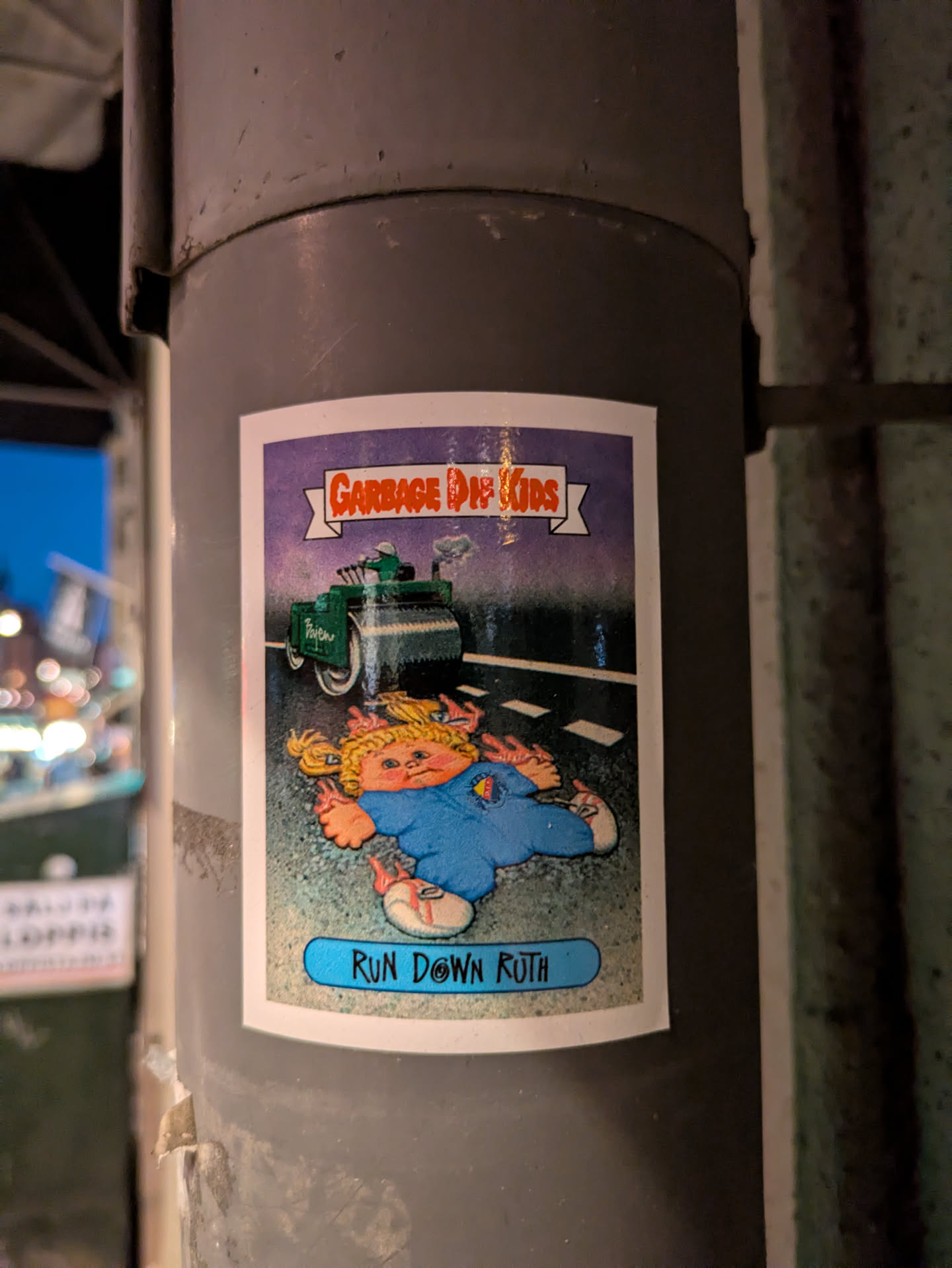 A rectangular sticker with a white border, depicting a scene from the 'Garbage Pail Kids' series. The central image features 'Run Down Ruth', a young girl with blonde pigtails and a blue jumpsuit, lying flattened on a dark grey road or pavement. Her limbs are splayed out, with red/pink squiggly lines suggesting gore. Behind her, a green steamroller, labeled 'Patches', is moving away, leaving a small cloud of smoke. The background is a dark purple sky. At the top, a white banner with orange, blocky letters reads 'GARBAGE PAIL KIDS'. A light blue banner at the bottom contains the name 'RUN DOWN RUTH' in black text. The art style is characteristic of GPK, featuring exaggerated, grotesque cartooning with vibrant colors. The sticker shows some minor wear, with slight peeling at the bottom left.