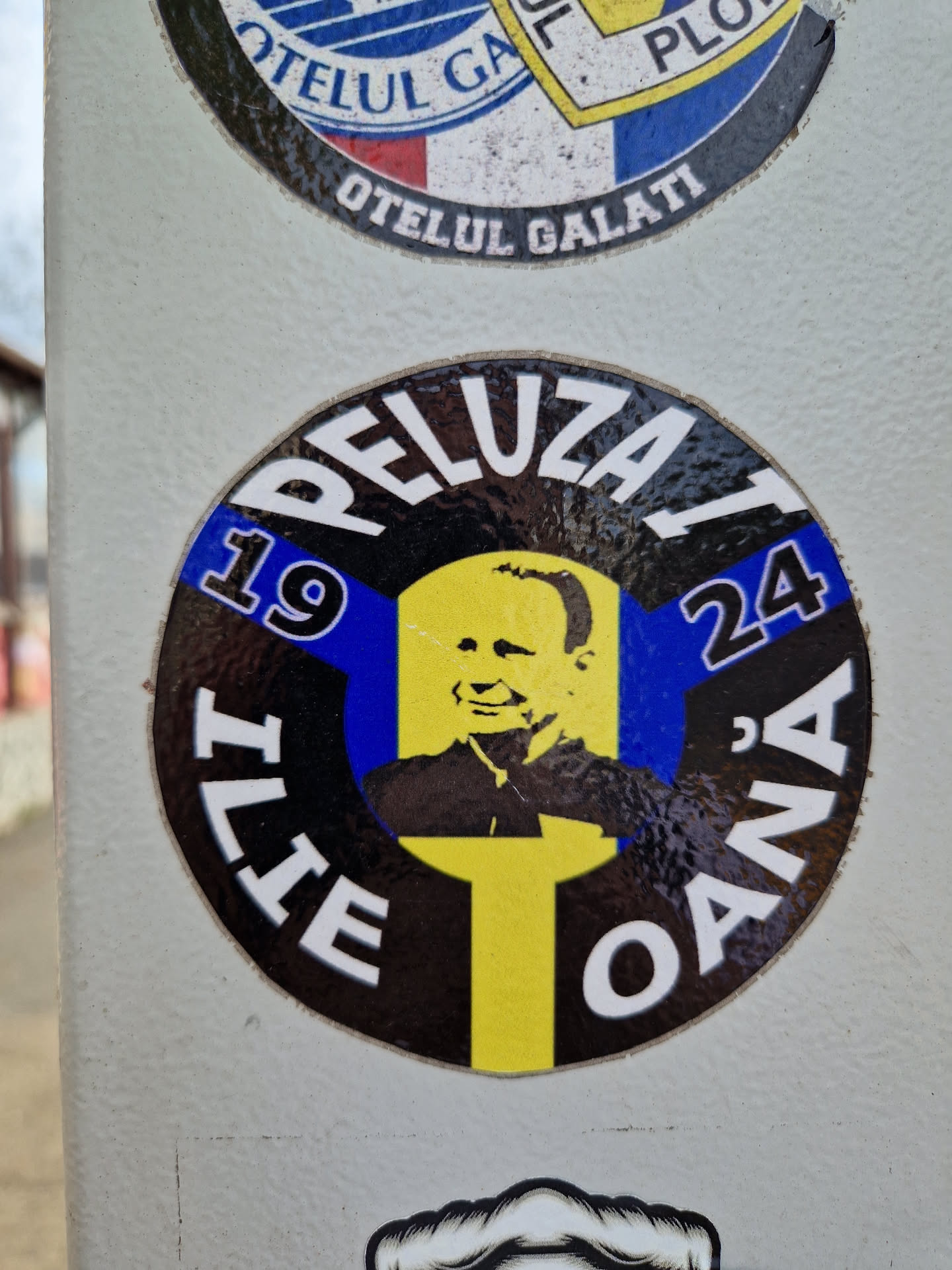 Round sticker with a black background and yellow and blue accents. It features a portrait of Ilie Oana and the text 'Peluza 19 Ilie Oana 24'.