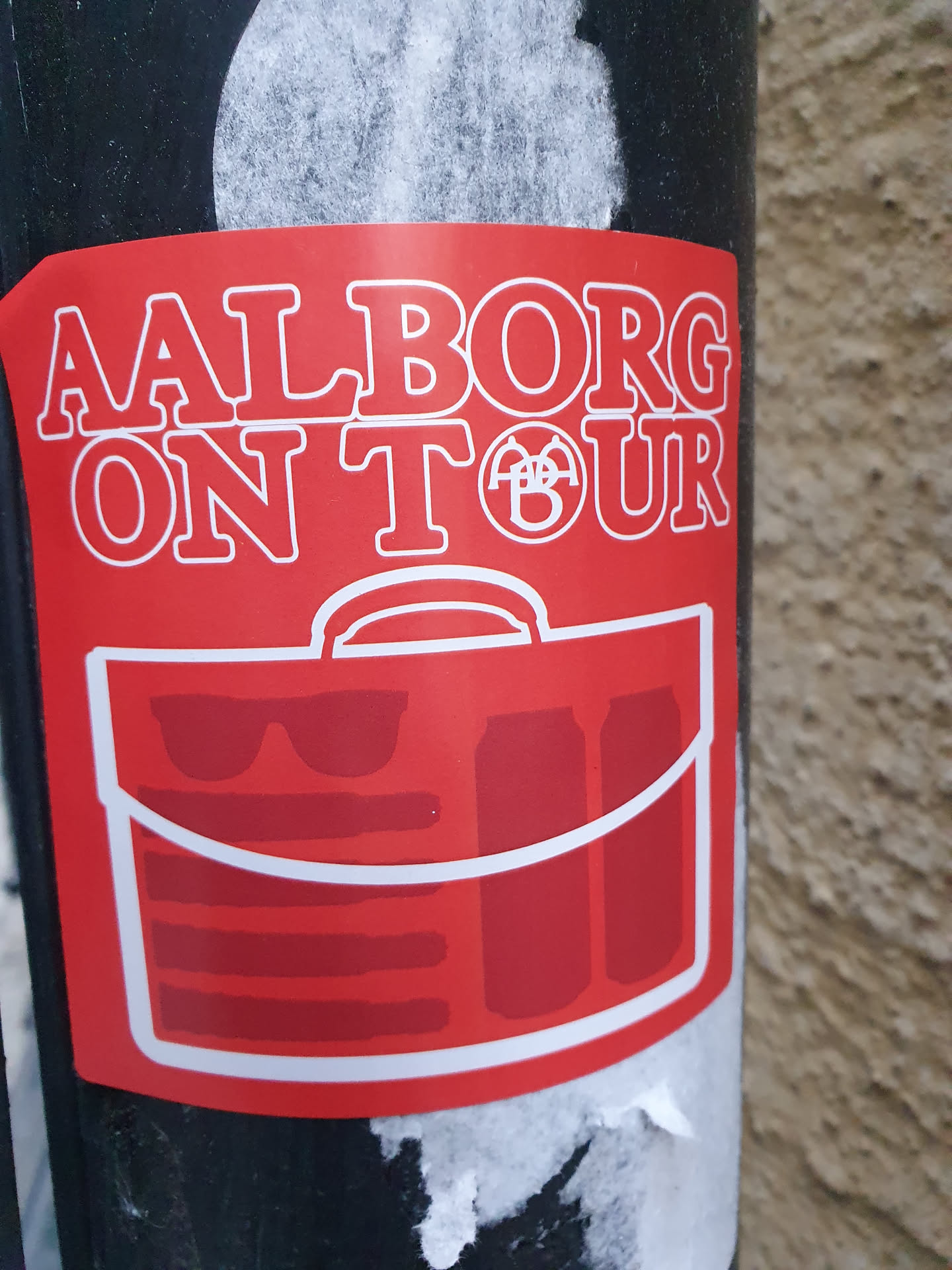 Red sticker with the text "Aalborg On Tour" and an illustration of a travel bag with sunglasses and cans inside. 