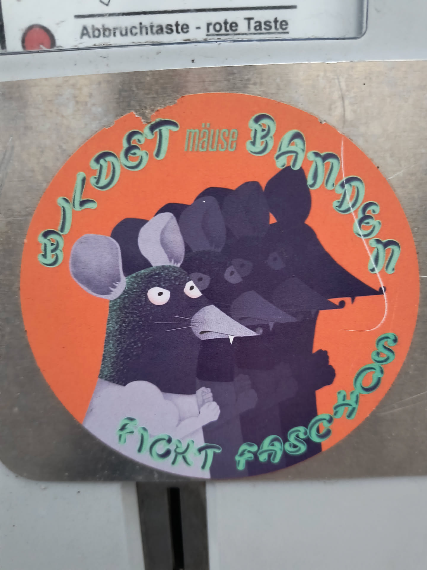 A circular sticker with a vibrant orange background featuring a group of stylized mice. The most prominent mouse on the left has a dark purple body with a stippled texture, lighter grey ears, and light grey forearms forming fists. It has white, wide-open eyes with small pupils and two pointed white fangs. Behind it, three other mice are depicted as darker purple silhouettes, suggesting a collective or 'gang'. The text 'BILDET mäuse BANDEN' is arranged along the top curve, and 'FICKT FASCHOS' along the bottom curve, both in light green with a dark outline. The sticker shows signs of wear, including a slight tear at the top edge.