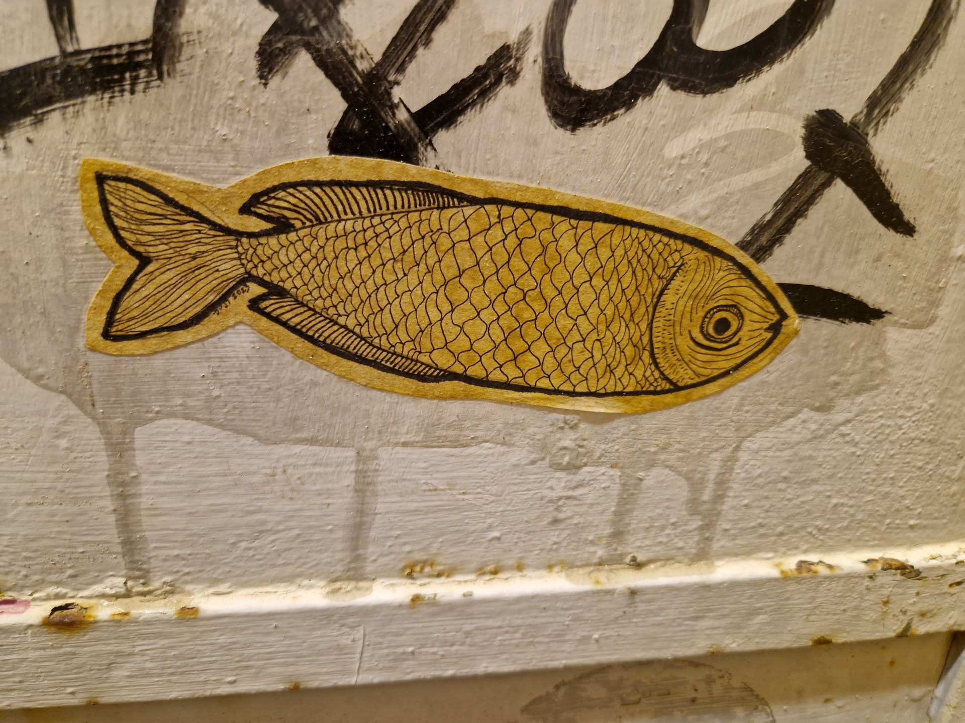 A yellow sticker with a hand-drawn illustration of a fish. The fish is detailed with scales and fins. The sticker is attached to a wall with some graffiti in the background.