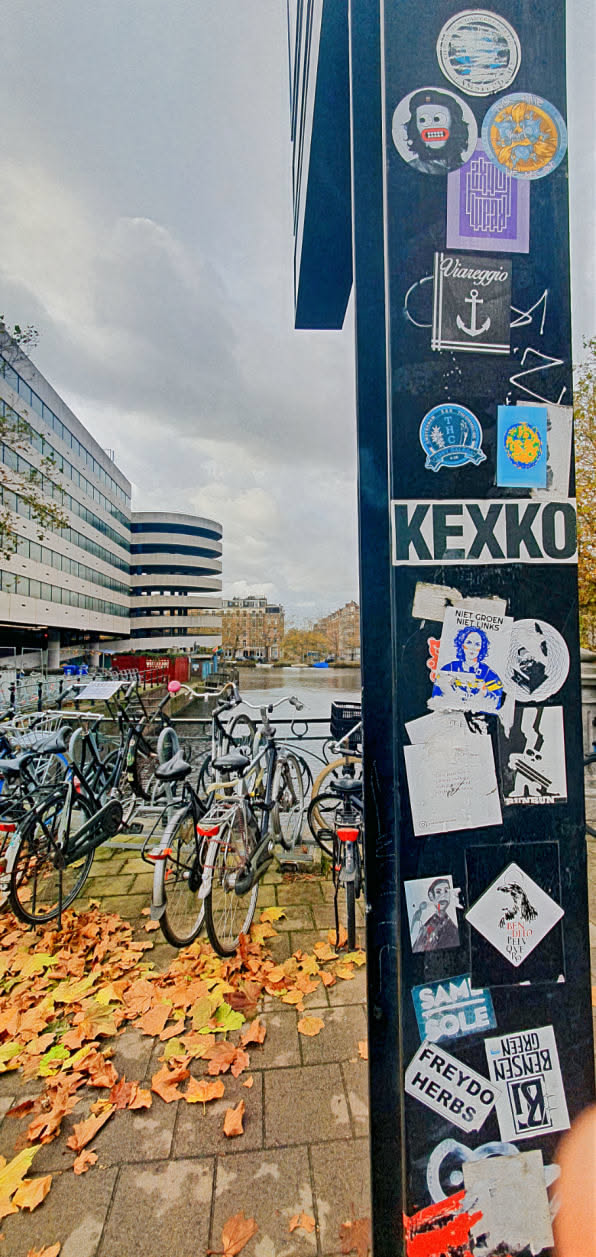 A black pole in Amsterdam is covered in stickers, including one for KEXKO, Freydo Herbs, and others. Bicycles are parked nearby, and fallen leaves cover the ground.