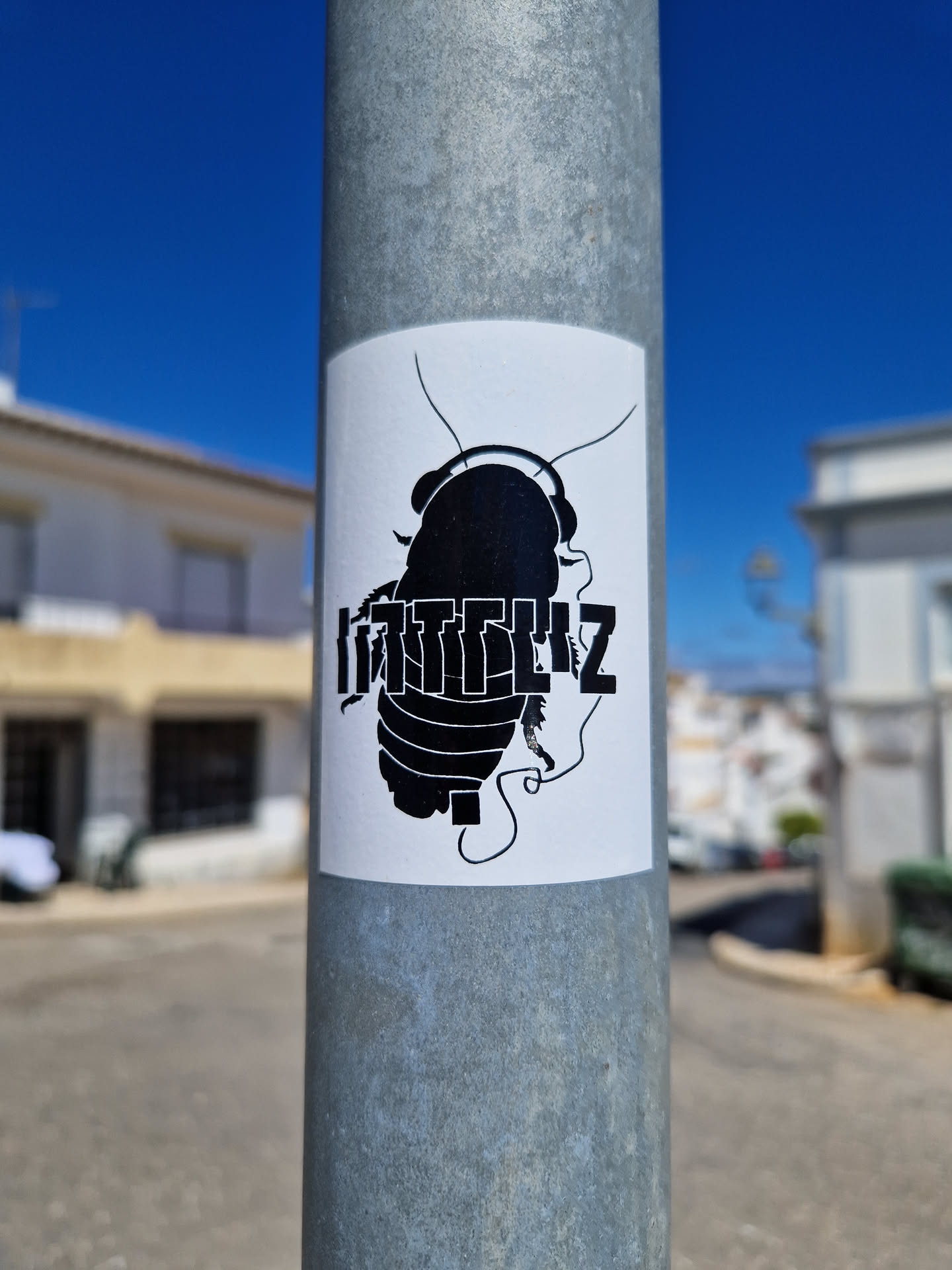 A black and white sticker of a cockroach wearing headphones. The text "KITTLEZ" is prominently displayed below the image of the cockroach. The sticker is affixed to a metal pole.