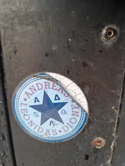 This circular sticker is cracked and peeling. It features a central five-pointed star with symbols in each point.  The text "Leonidas Andreas Dione" is written in a circular pattern around the star.