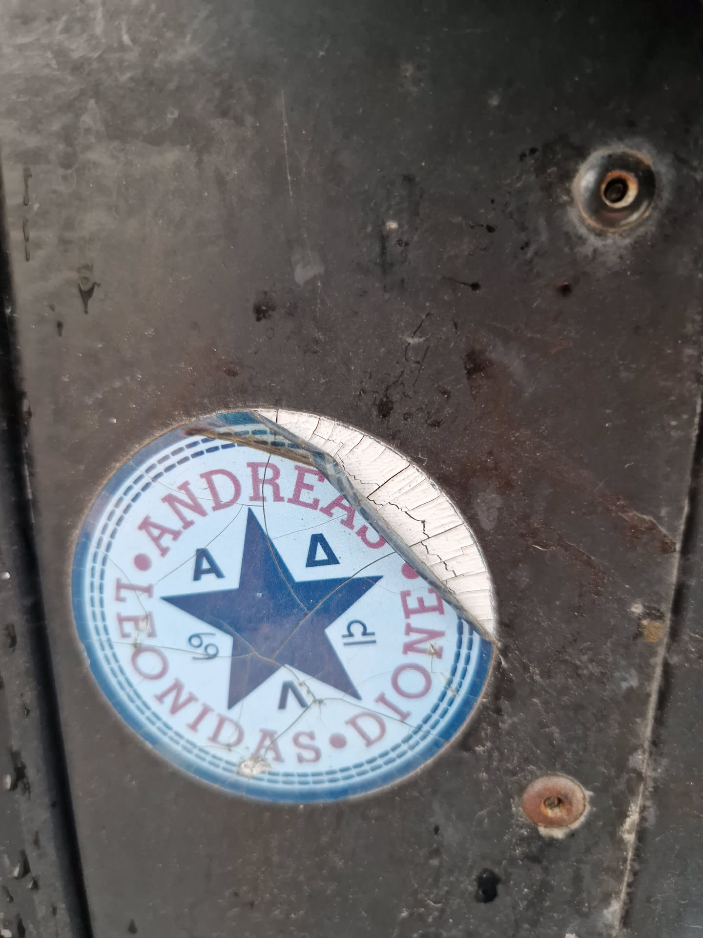 This circular sticker is cracked and peeling. It features a central five-pointed star with symbols in each point.  The text "Leonidas Andreas Dione" is written in a circular pattern around the star.