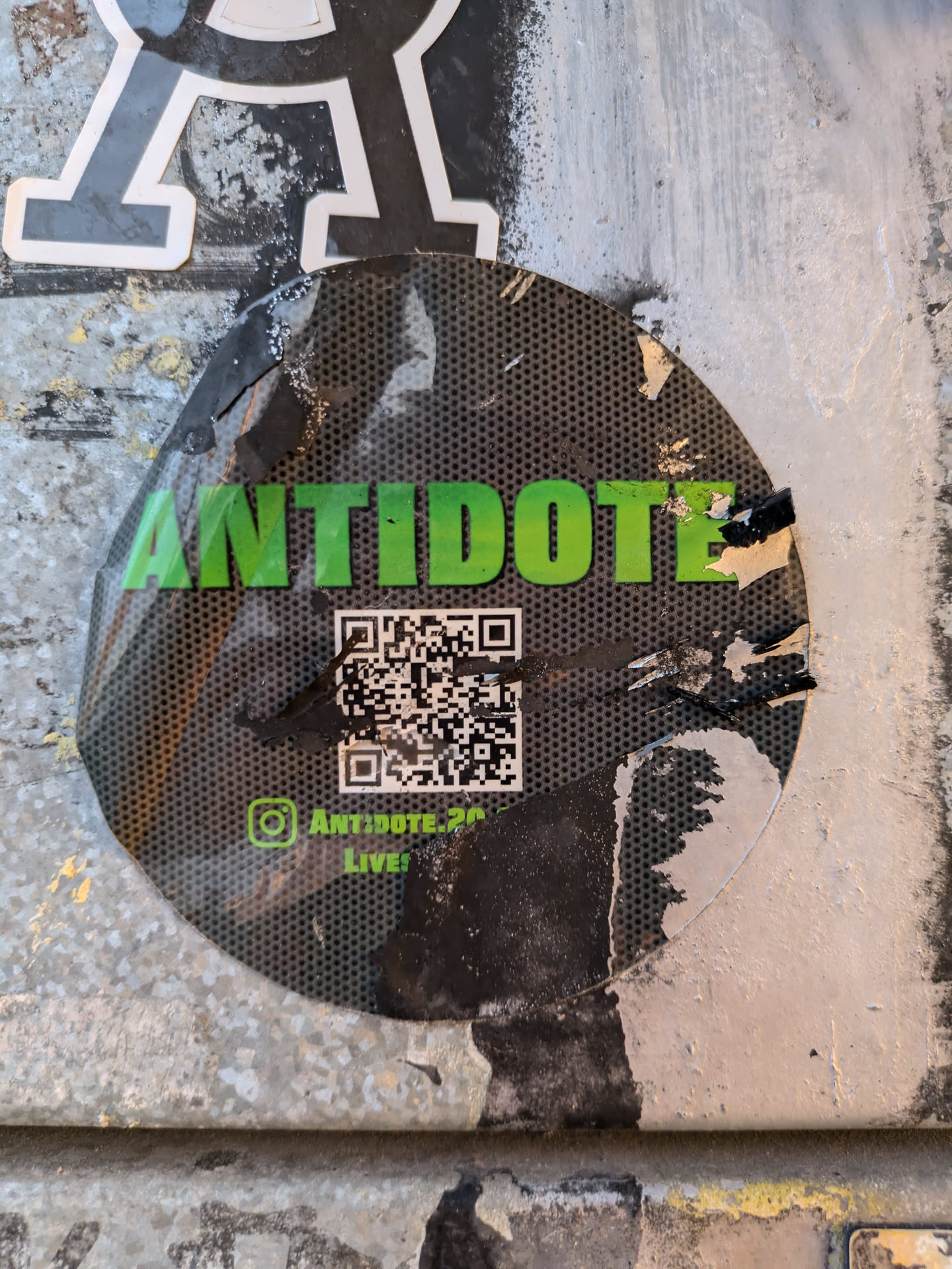 A round black and green sticker with the word 'ANTIDOTE' in large green letters. Below the text, there is a QR code and Instagram handle @antidote_26. The sticker is slightly damaged and peeling from the surface. It is attached to a wall with other graffiti and stickers.